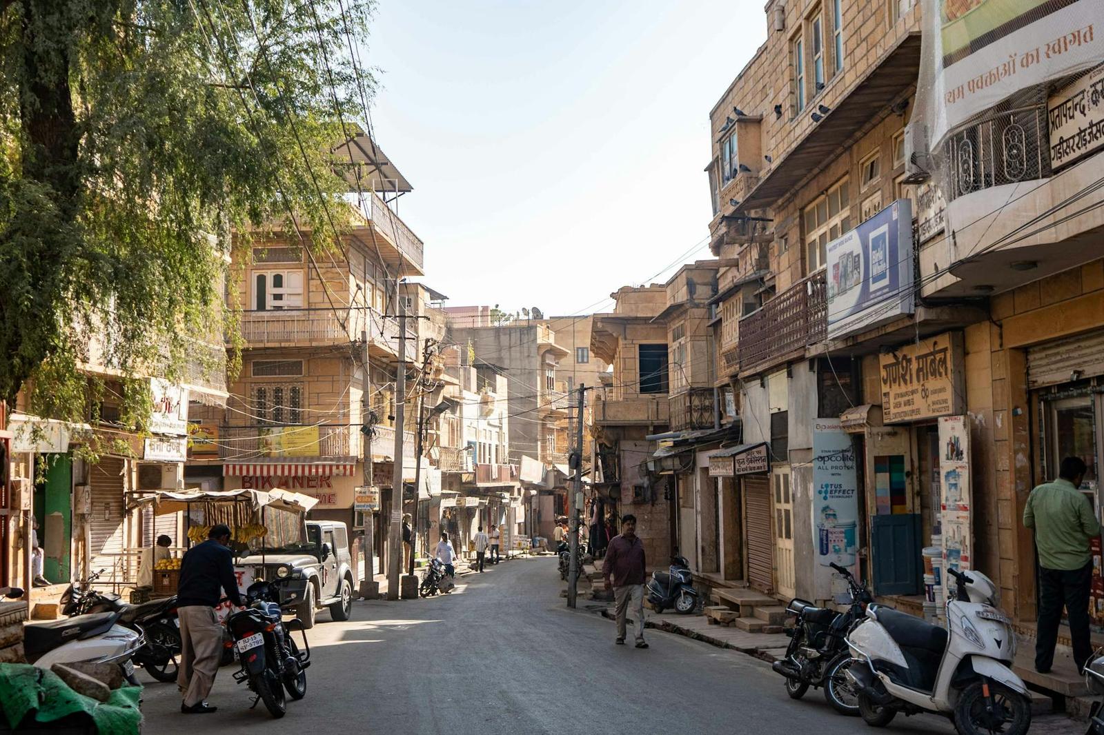 A quiet old-town street with sandstone buildings, parked scooters and a few pedestrians