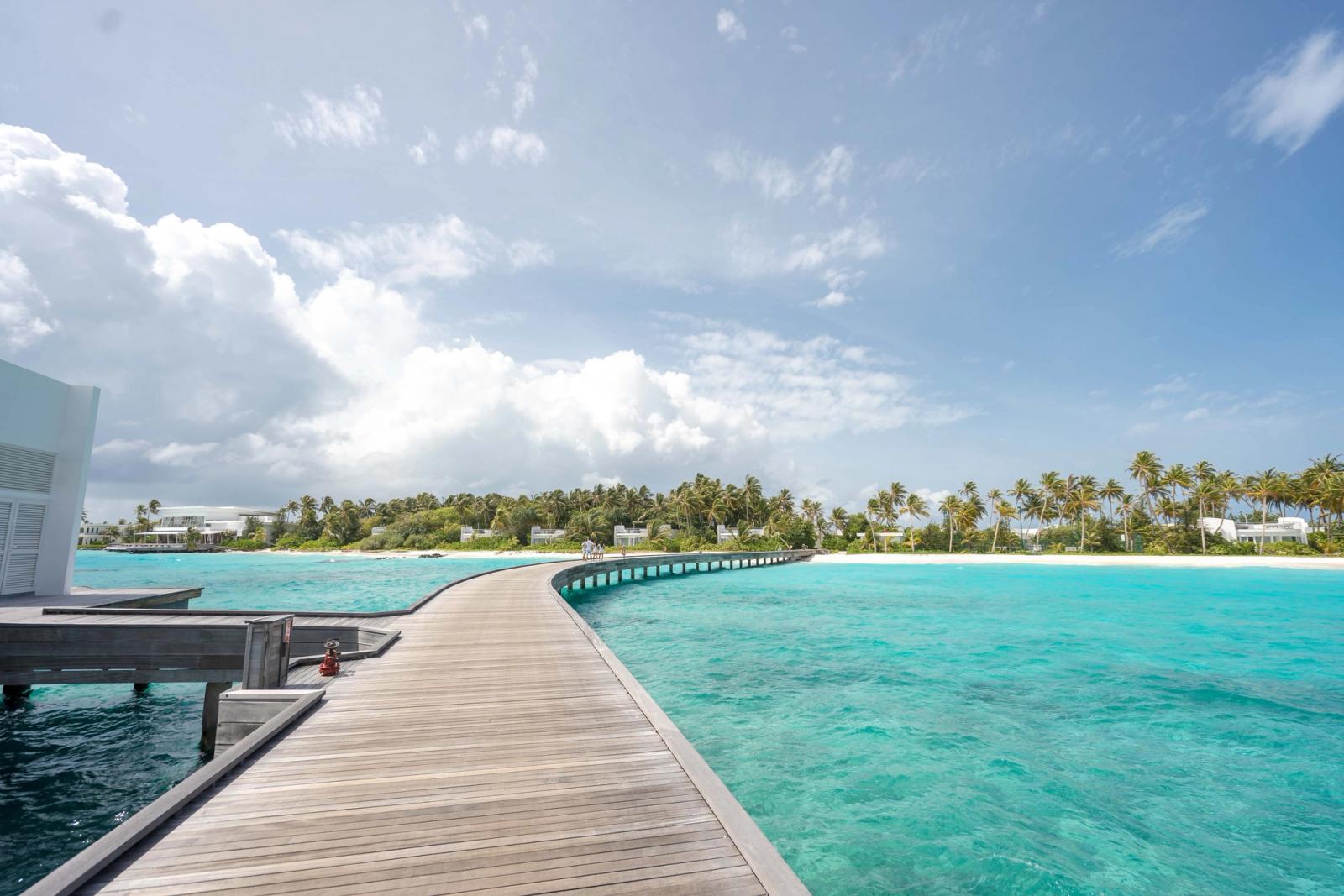 A wooden boardwalk stretching across a lagoon toward palm-lined shore