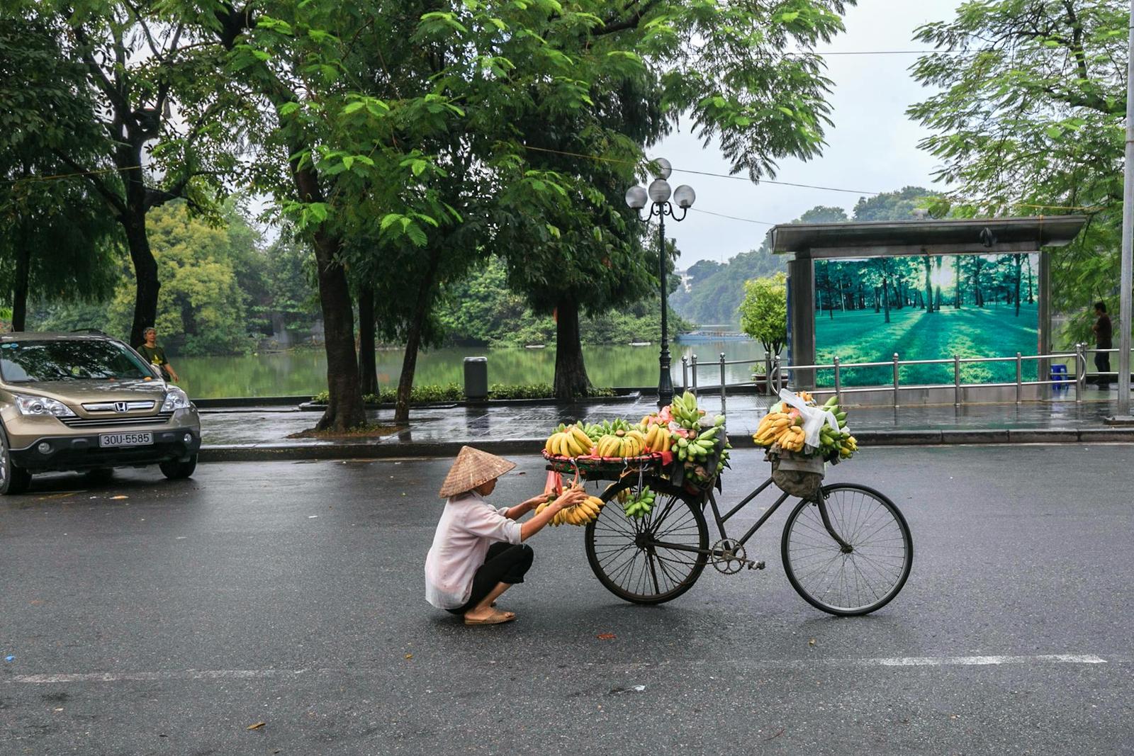 Street vendor crouching beside a bicycle loaded with baskets of fruit in the rain