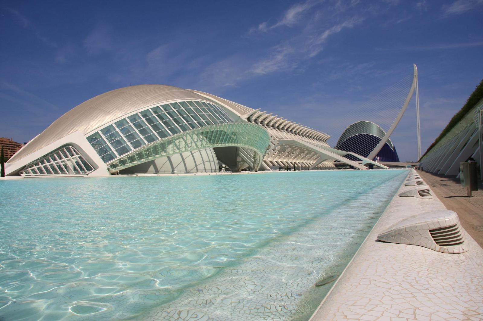 Curved glass and white concrete building beside a turquoise reflecting pool under blue sky