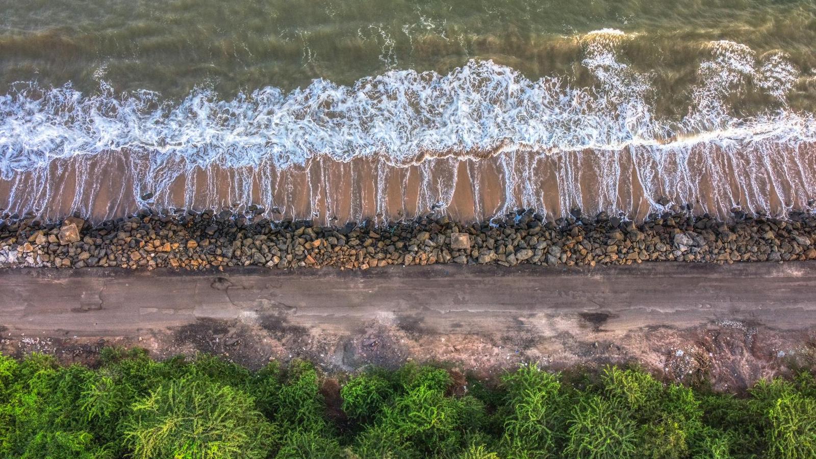Aerial view of waves breaking on a stone-lined shore beside green scrub