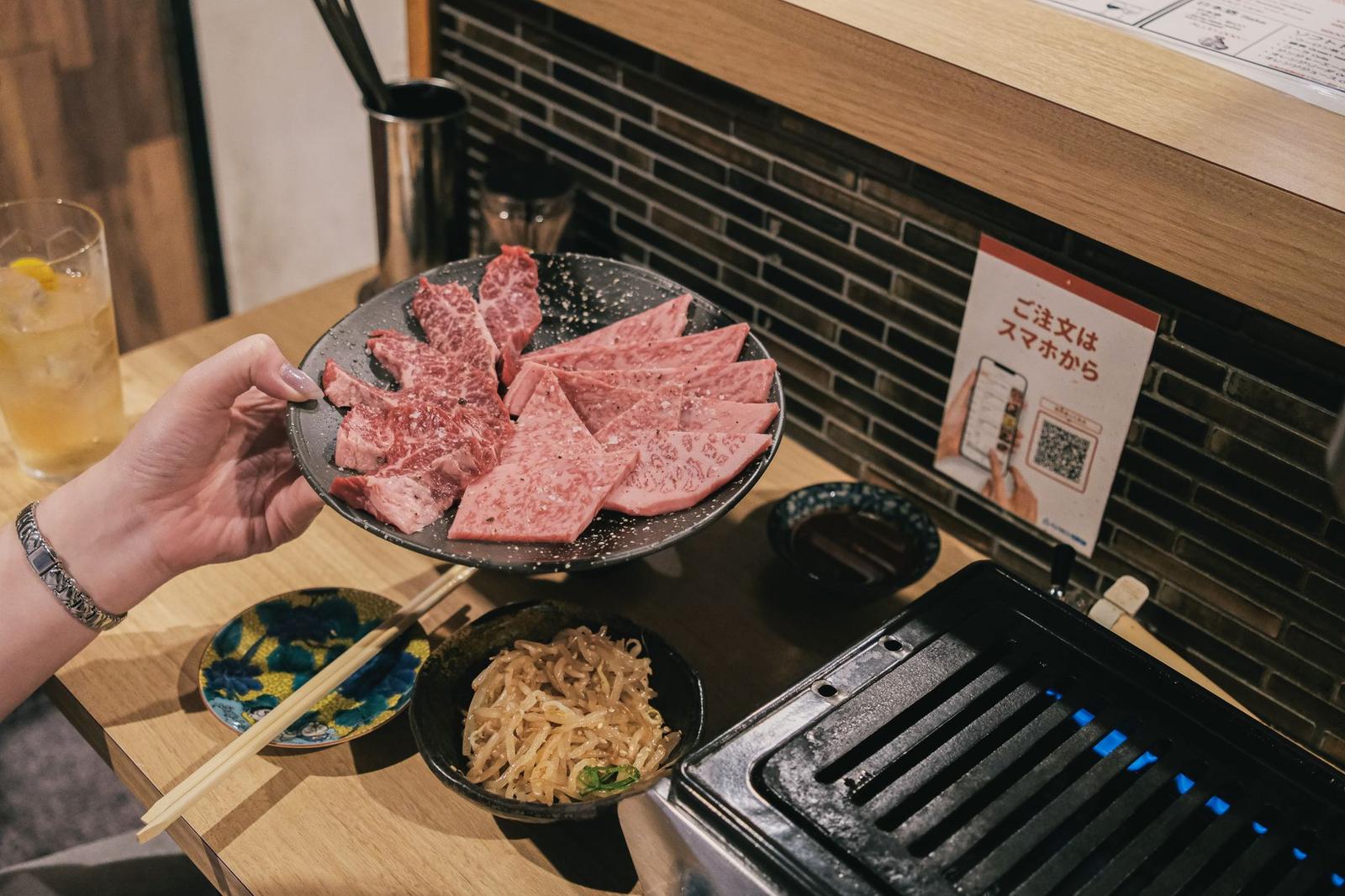 Hand with chopsticks lifting thinly sliced marbled beef from a platter beside side dishes