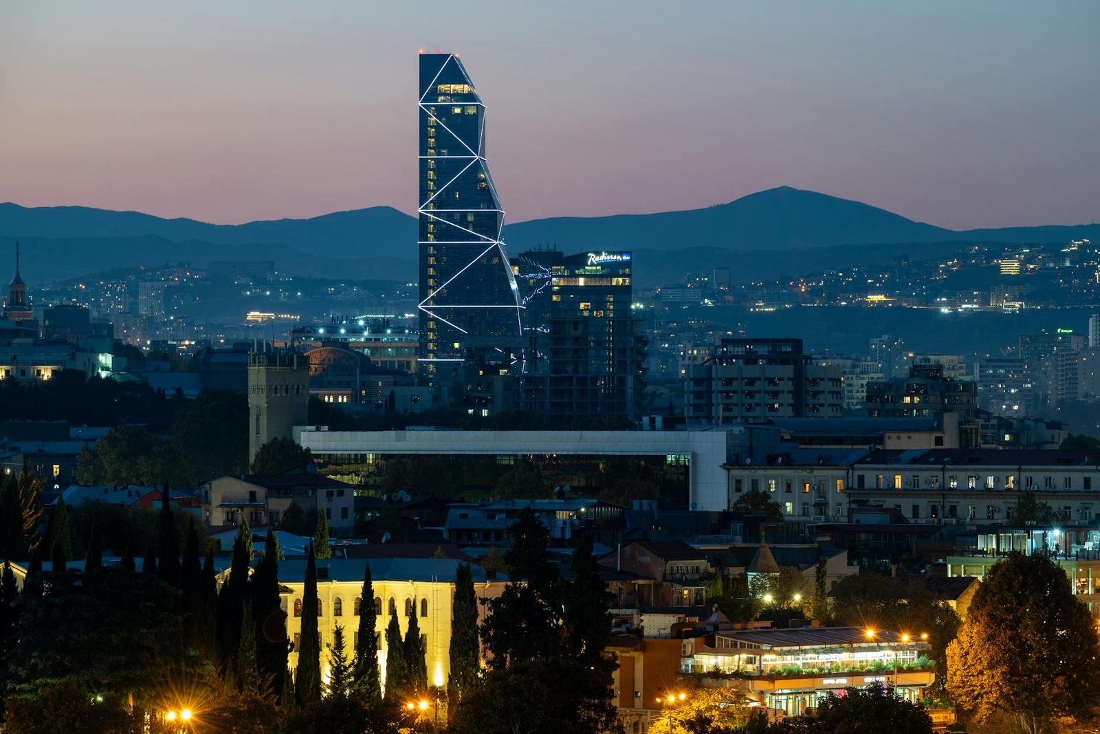 An angular glass skyscraper lit up at dusk with hills in the distance