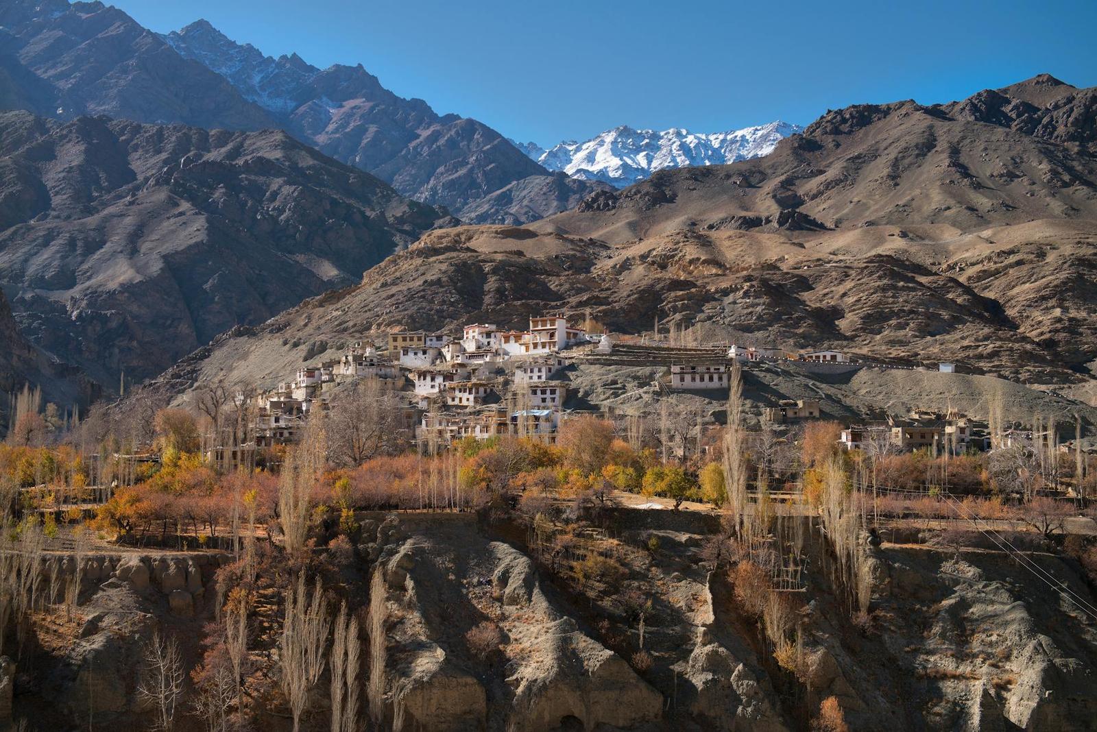 A village with autumn-colored trees set below jagged snow-capped mountains
