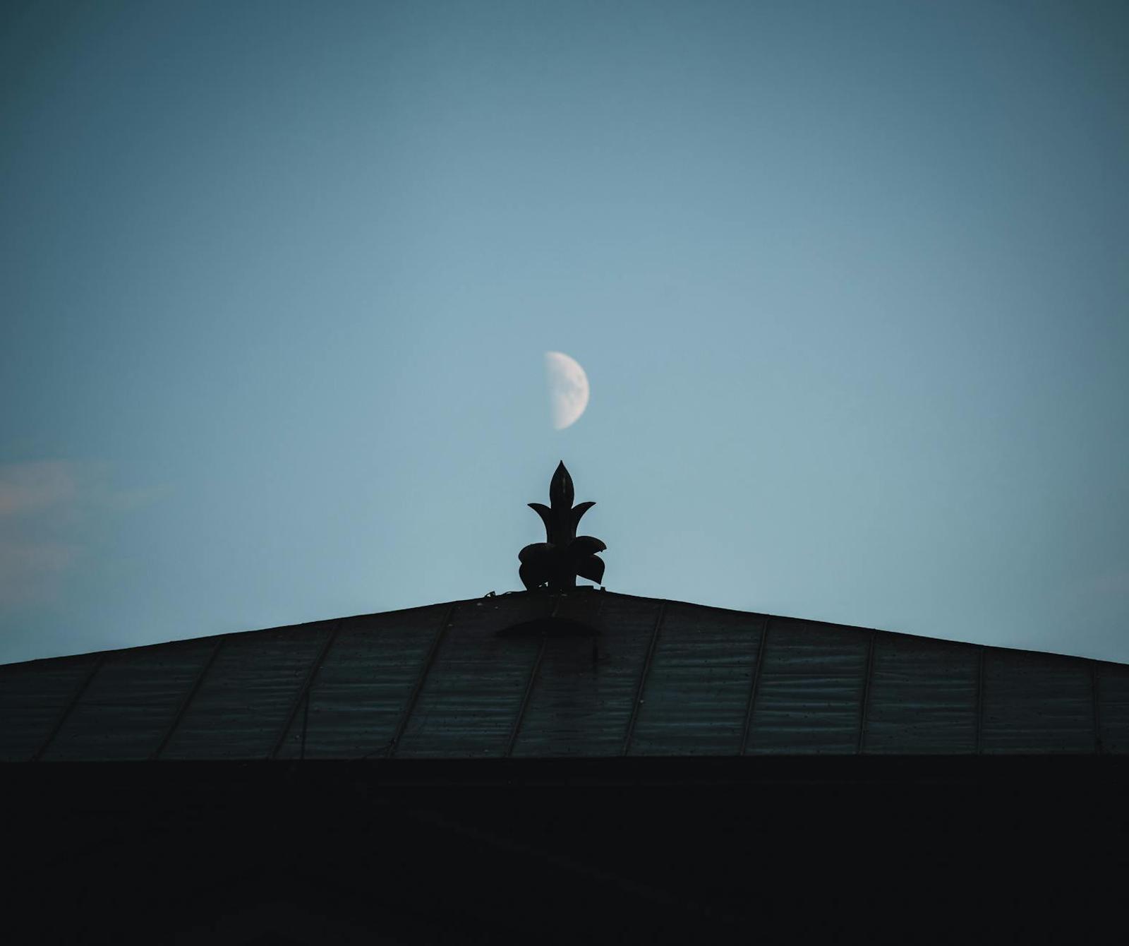 A carved rooftop finial silhouetted against a half moon at dusk