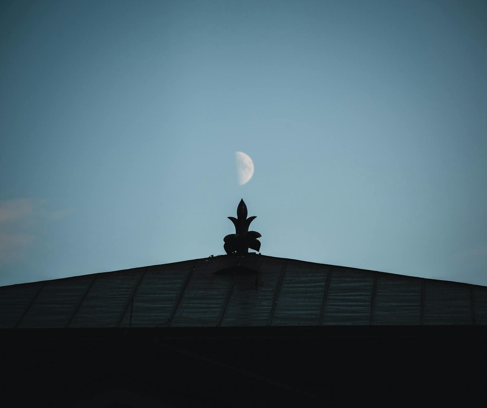 A carved rooftop finial silhouetted against a half moon at dusk
