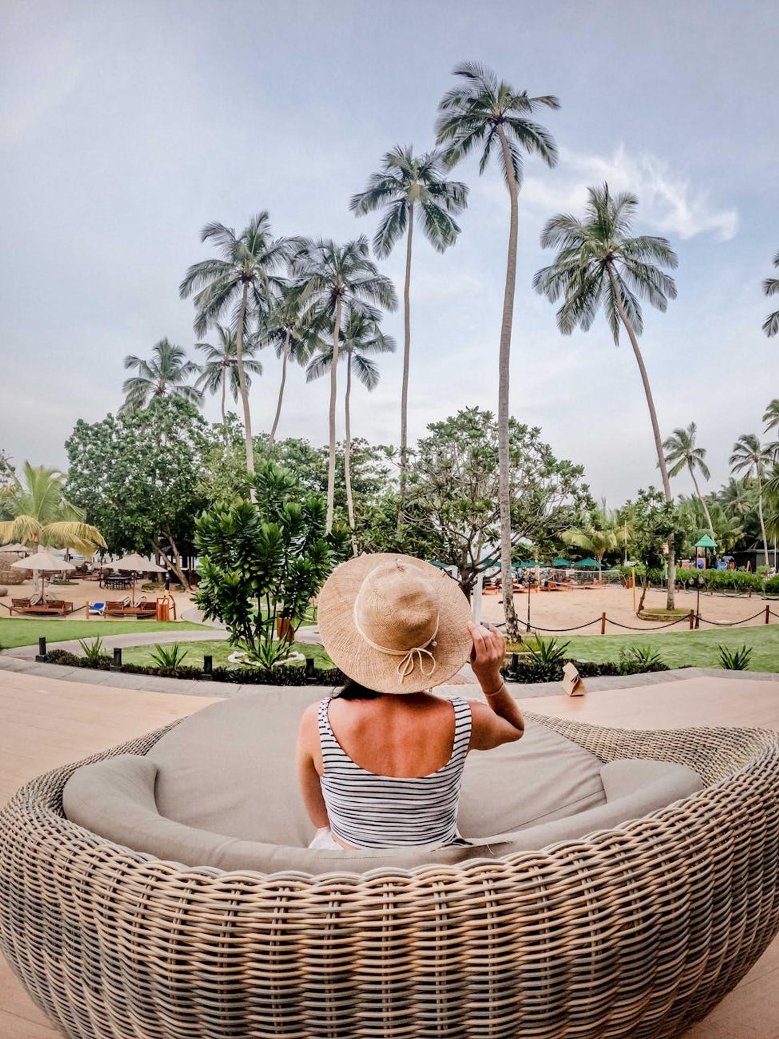 Woman in a sun hat seated on a wicker daybed among palm trees