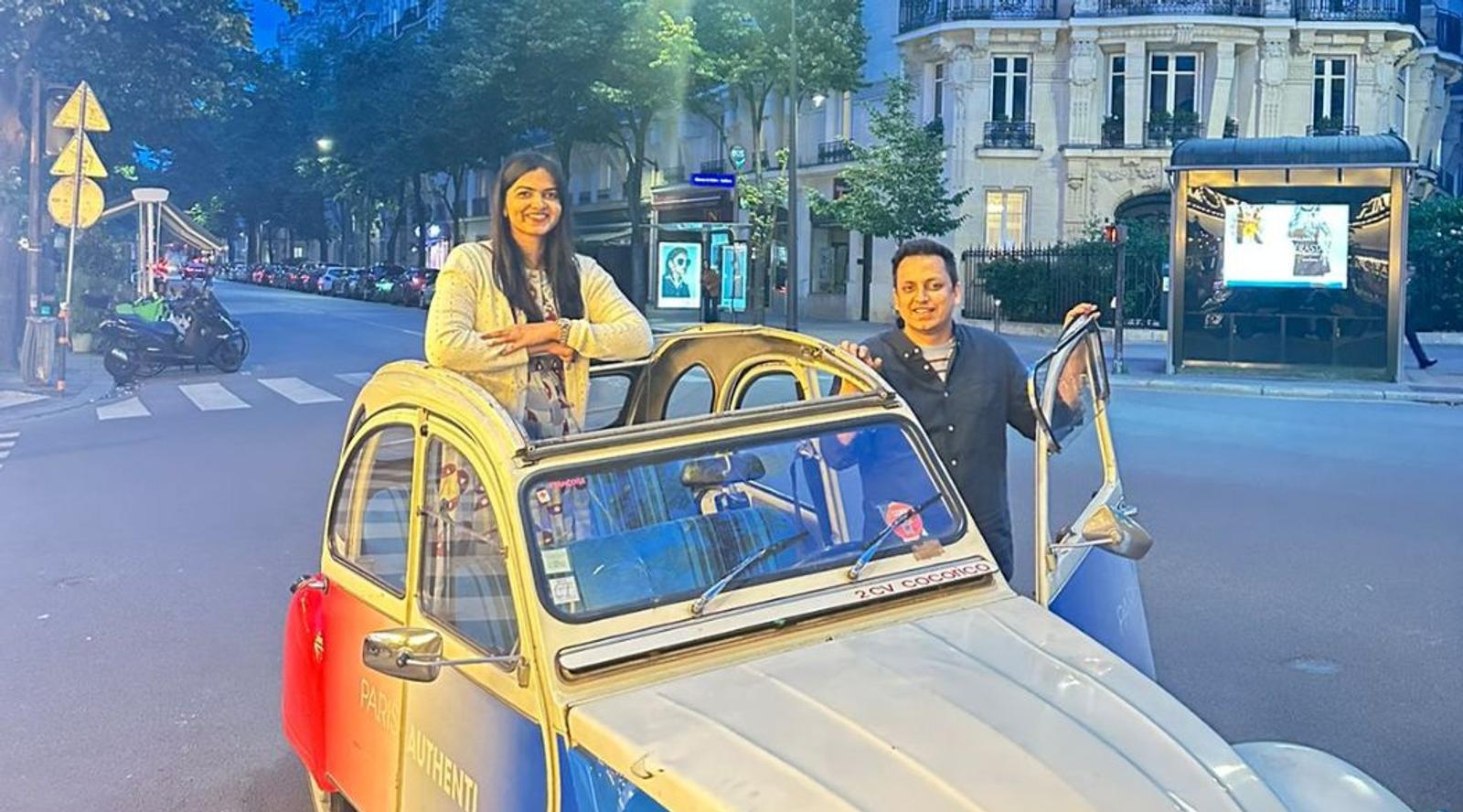 Two women posing with a vintage French car on a Paris street.