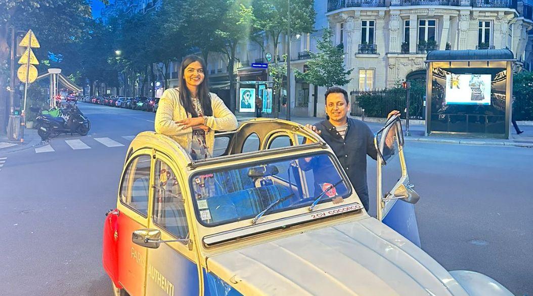 Two women posing with a vintage French car on a Paris street.