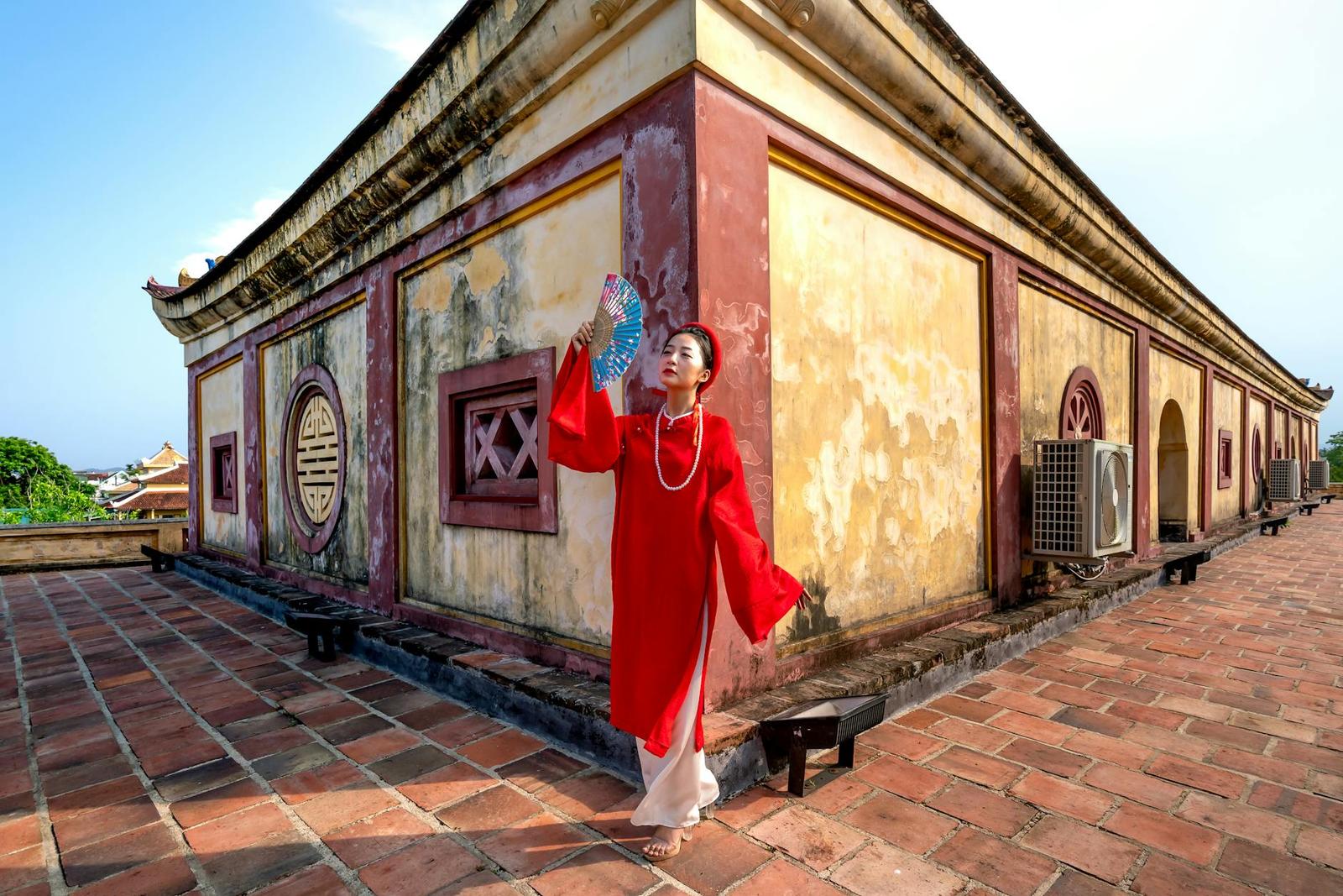 Woman in a red silk dress holding a fan beside a weathered ochre temple wall
