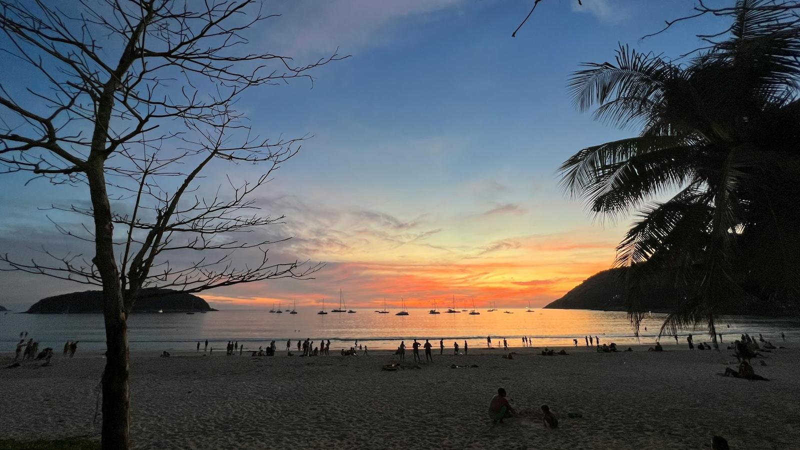 Silhouetted trees frame a crowded beach at sunset with anchored sailboats offshore