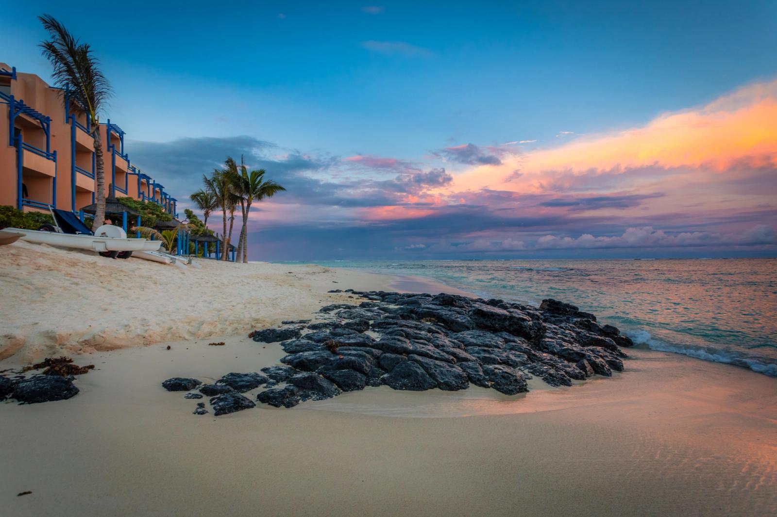 Beachfront resort buildings and palm trees at sunset beside dark rocks