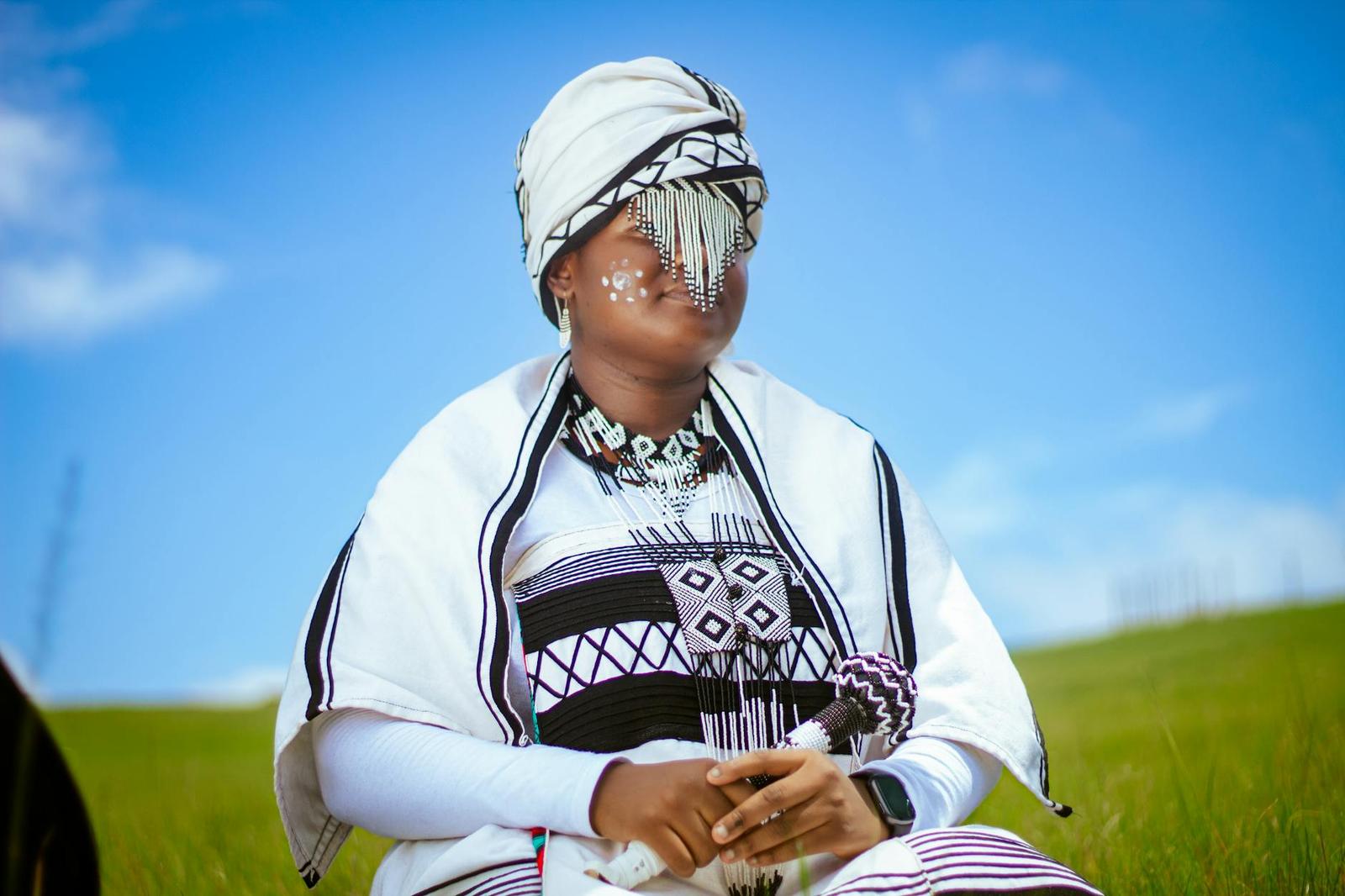 Woman in traditional white and black beaded ceremonial dress seated in a grassy field