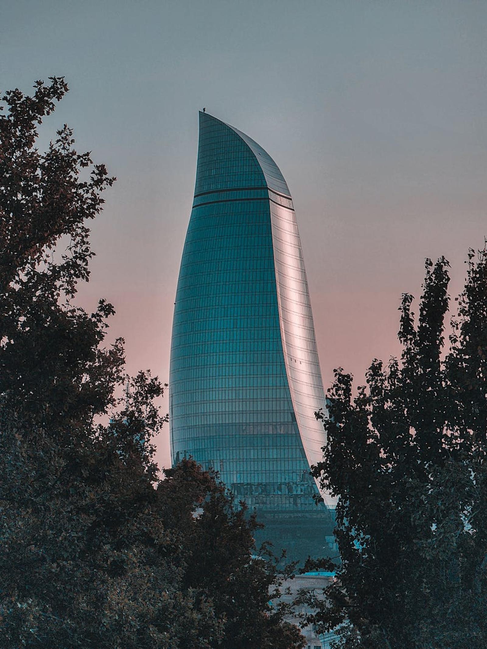 A sleek skyscraper in Baku cityscape at dusk, framed by trees highlighting urban architecture