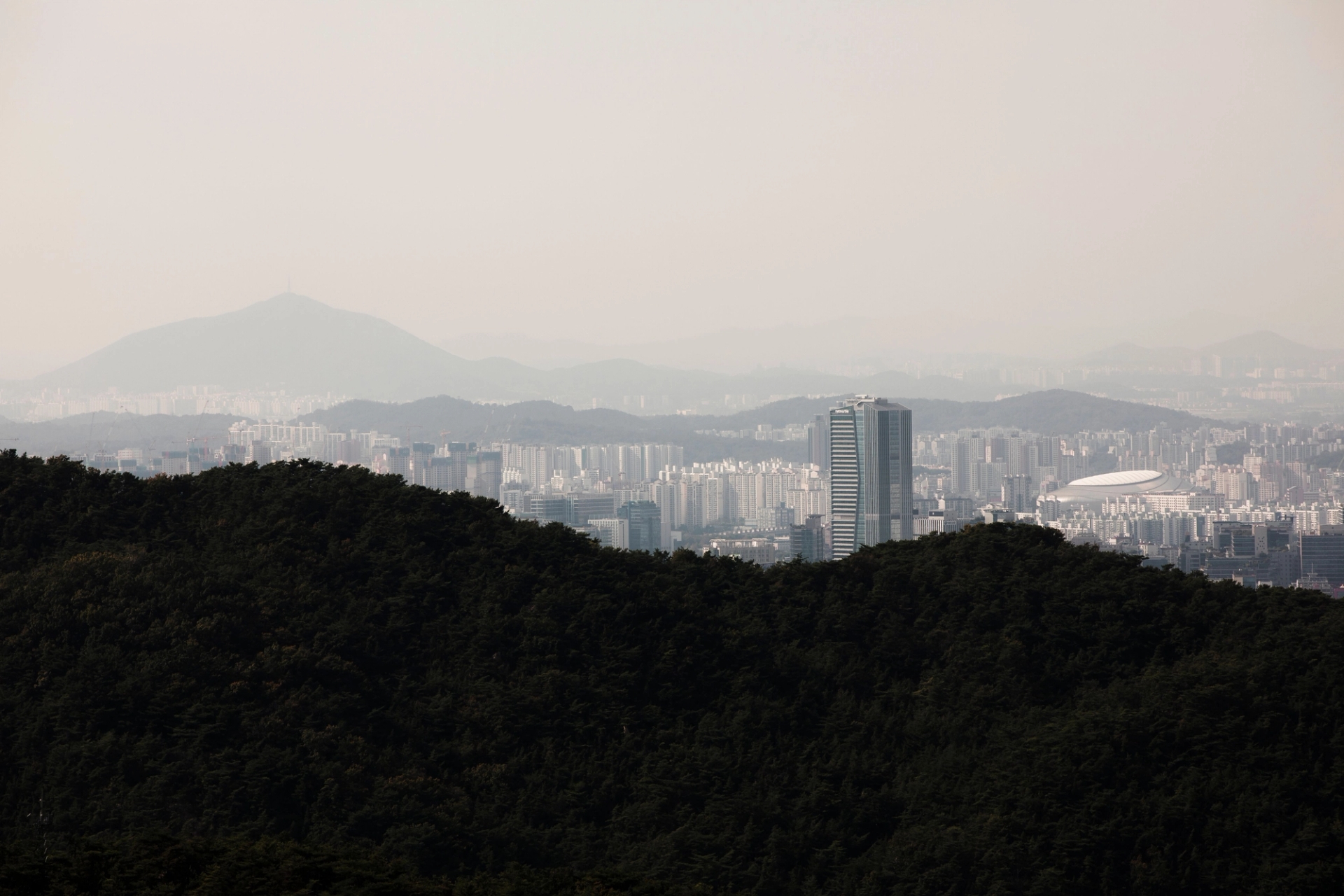 Aerial skyline of Seoul from forested hillsides, featuring distant mountains