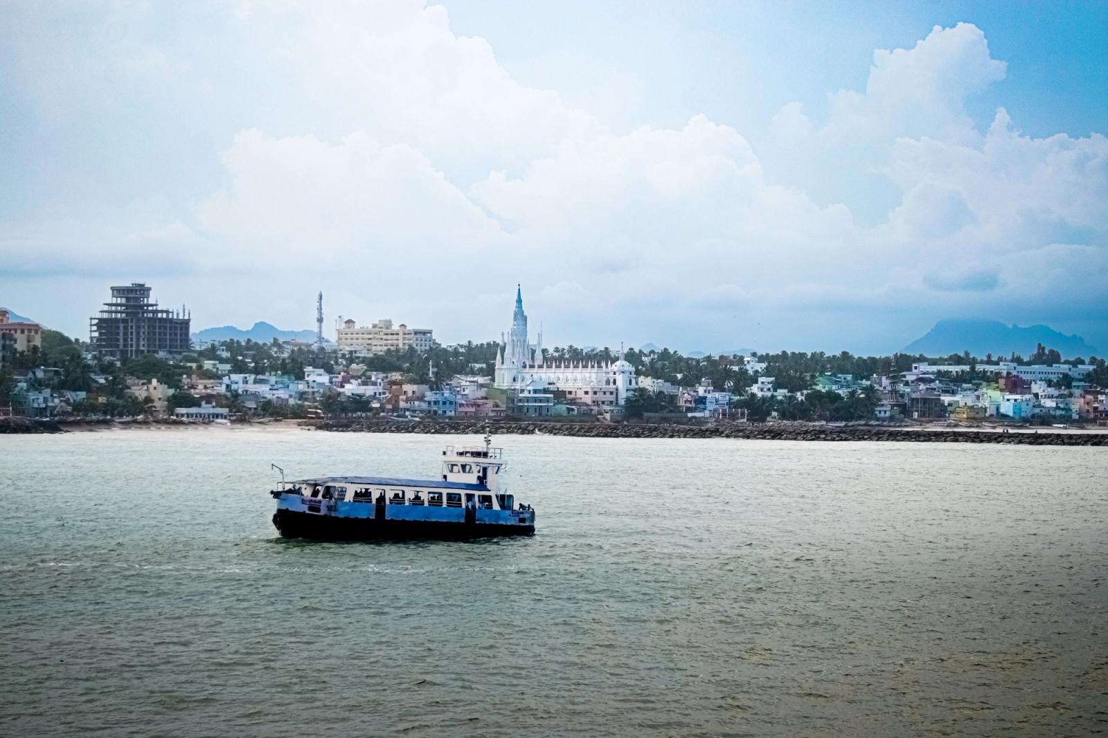 A passenger ship sails past the scenic waterfront of Kanyakumari, India, with urban landscapes in the background