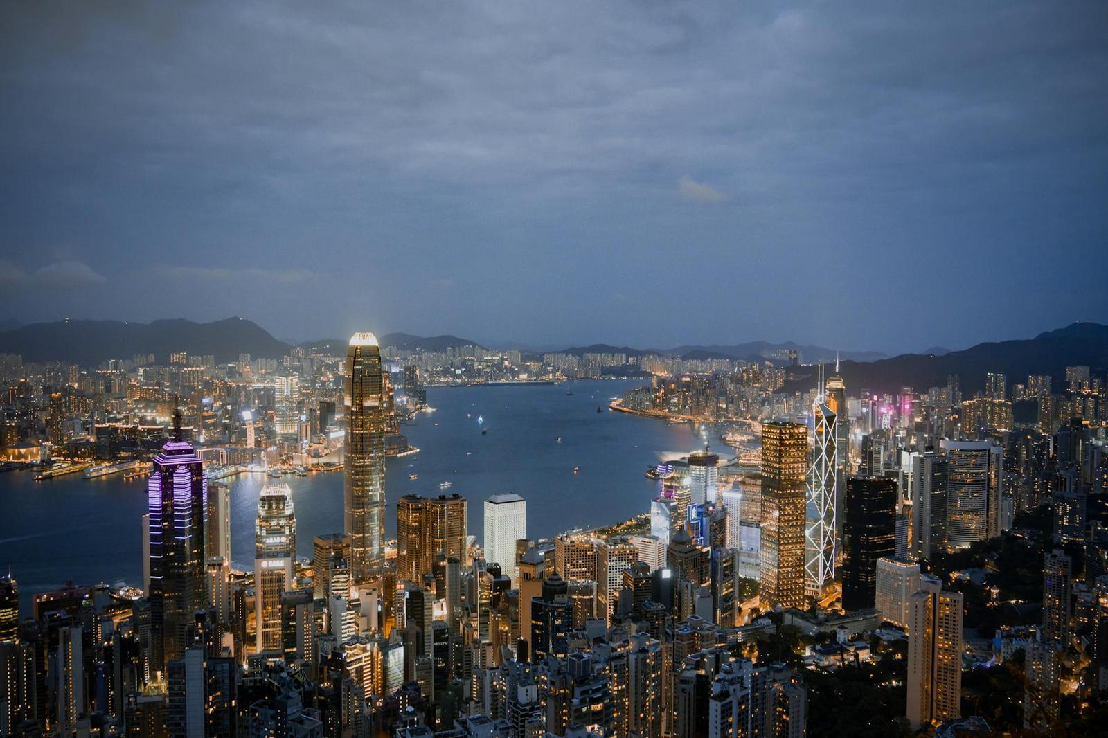 A harbourfront skyline of skyscrapers glowing at dusk beneath cloudy skies
