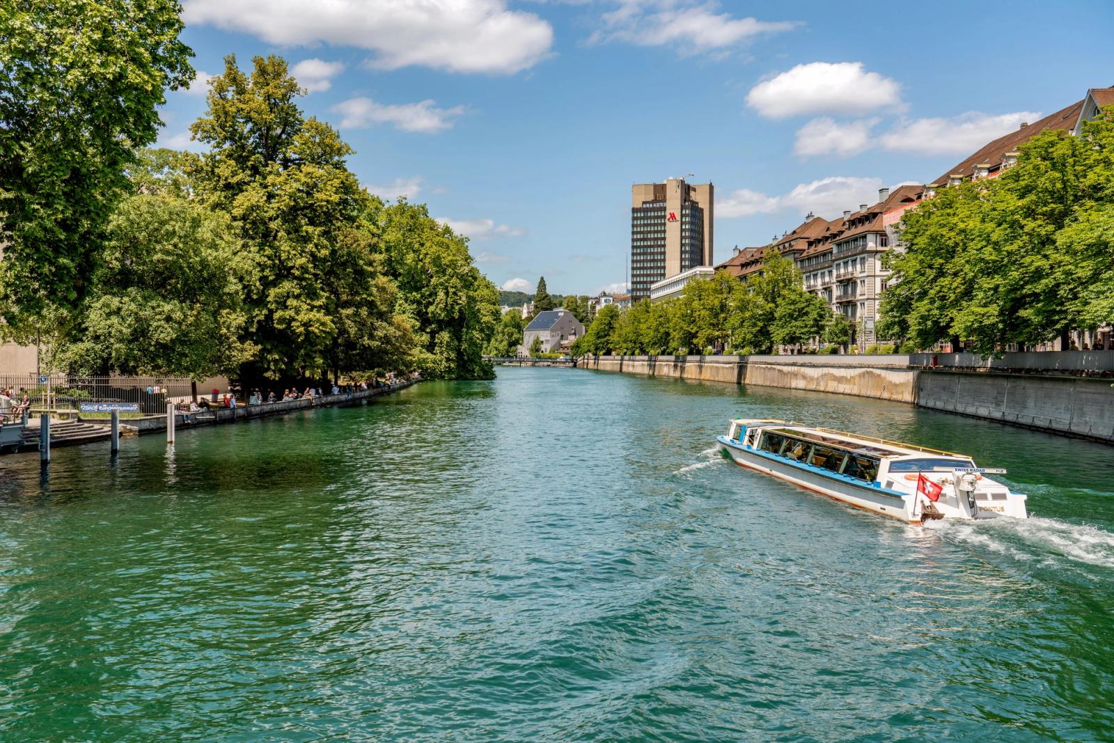 A tranquil view of a river in Zurich with a boat sailing under a blue, sunny sky