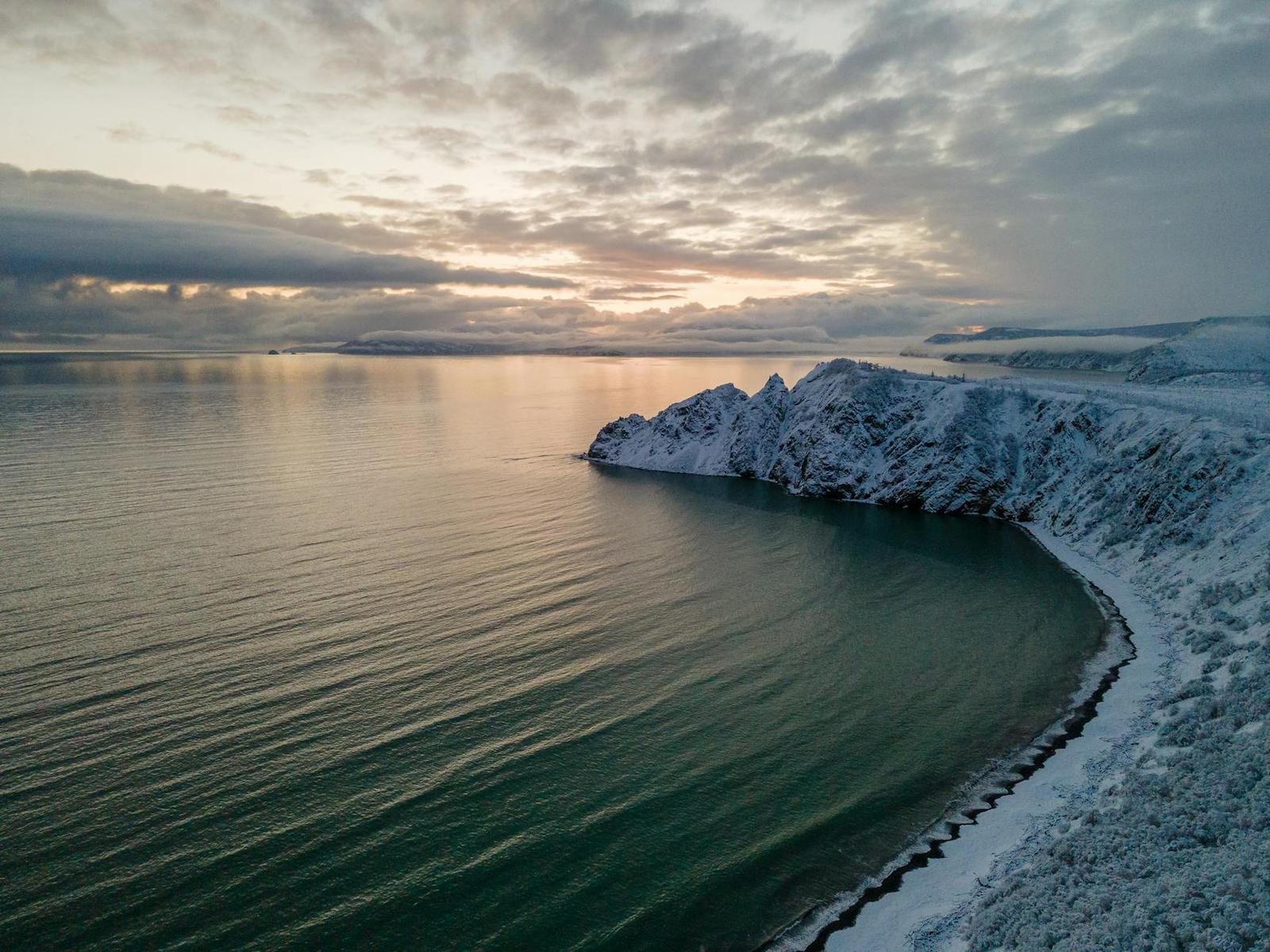 A snow-dusted headland curving into calm sea under a pale winter sunset