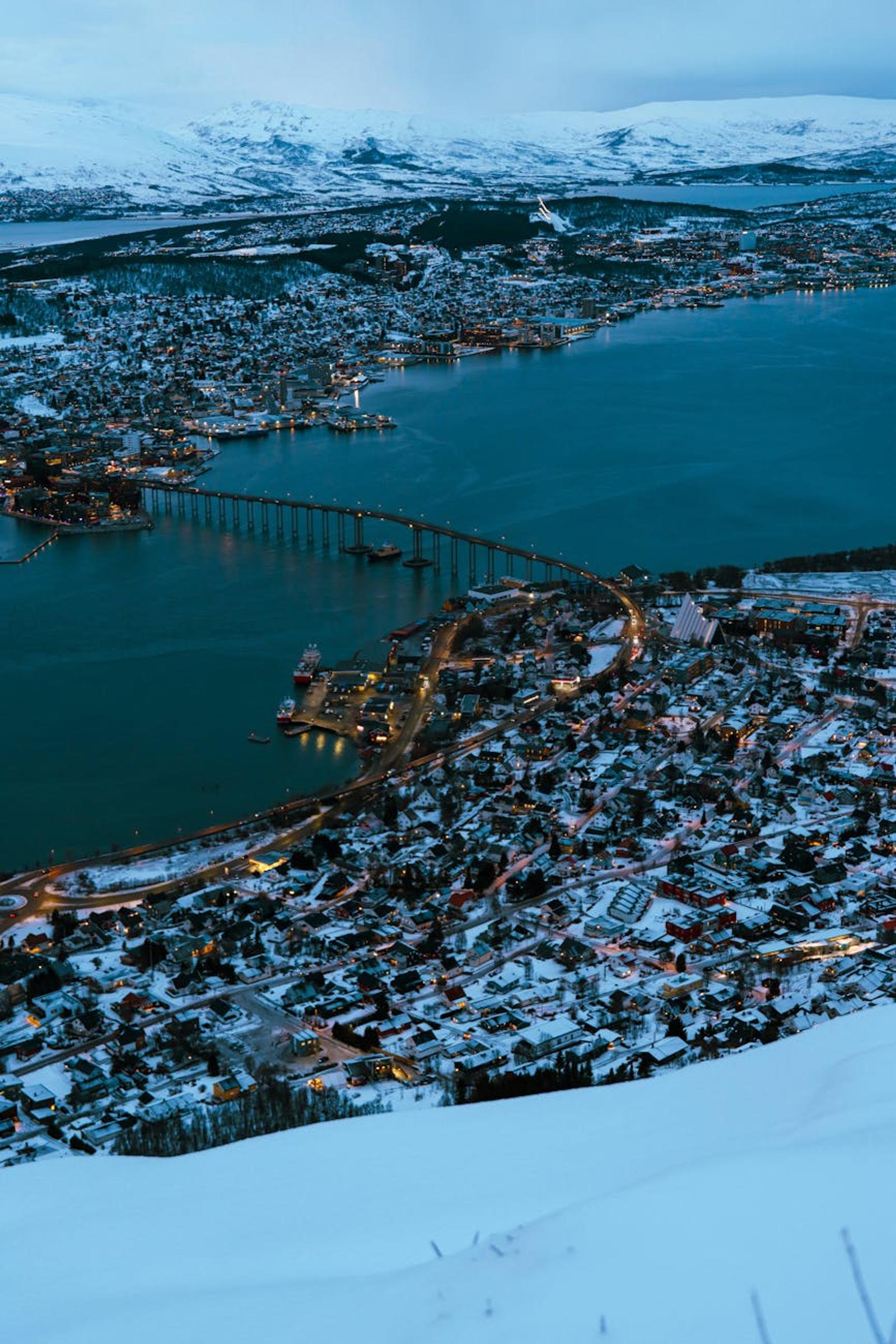 Aerial dusk view of a snow-covered fjord city linked by a long illuminated bridge
