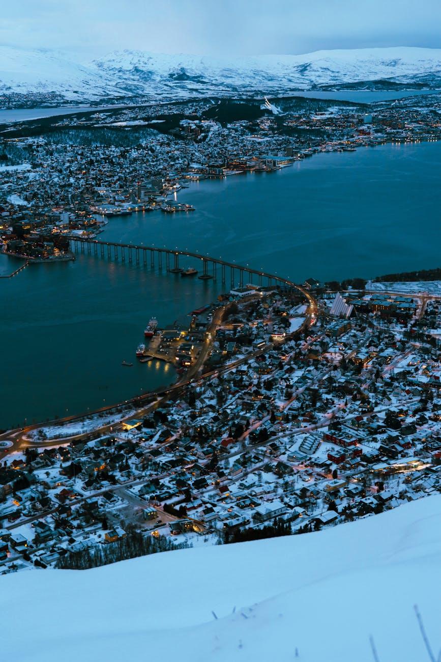 Aerial dusk view of a snow-covered fjord city linked by a long illuminated bridge