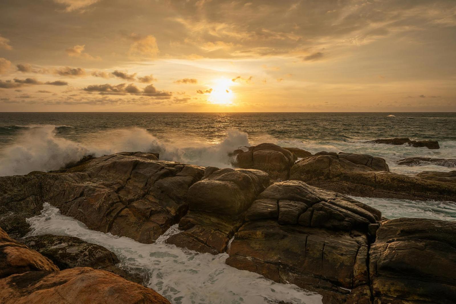 Waves breaking over dark coastal rocks under a golden sunset sky