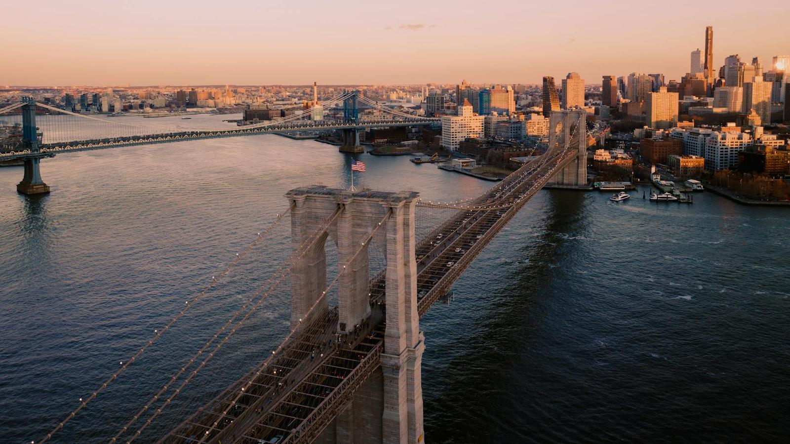 A stone suspension bridge crossing a wide river toward a city skyline at dusk