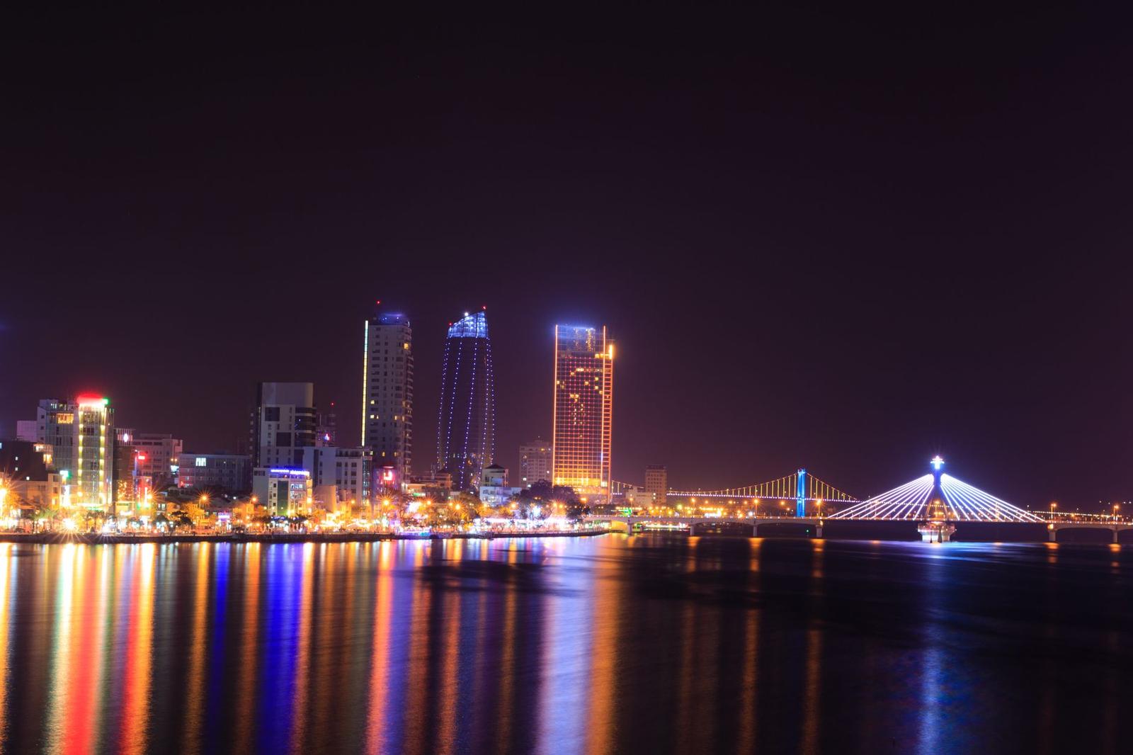 A city skyline reflected on the water with a curved illuminated bridge at night