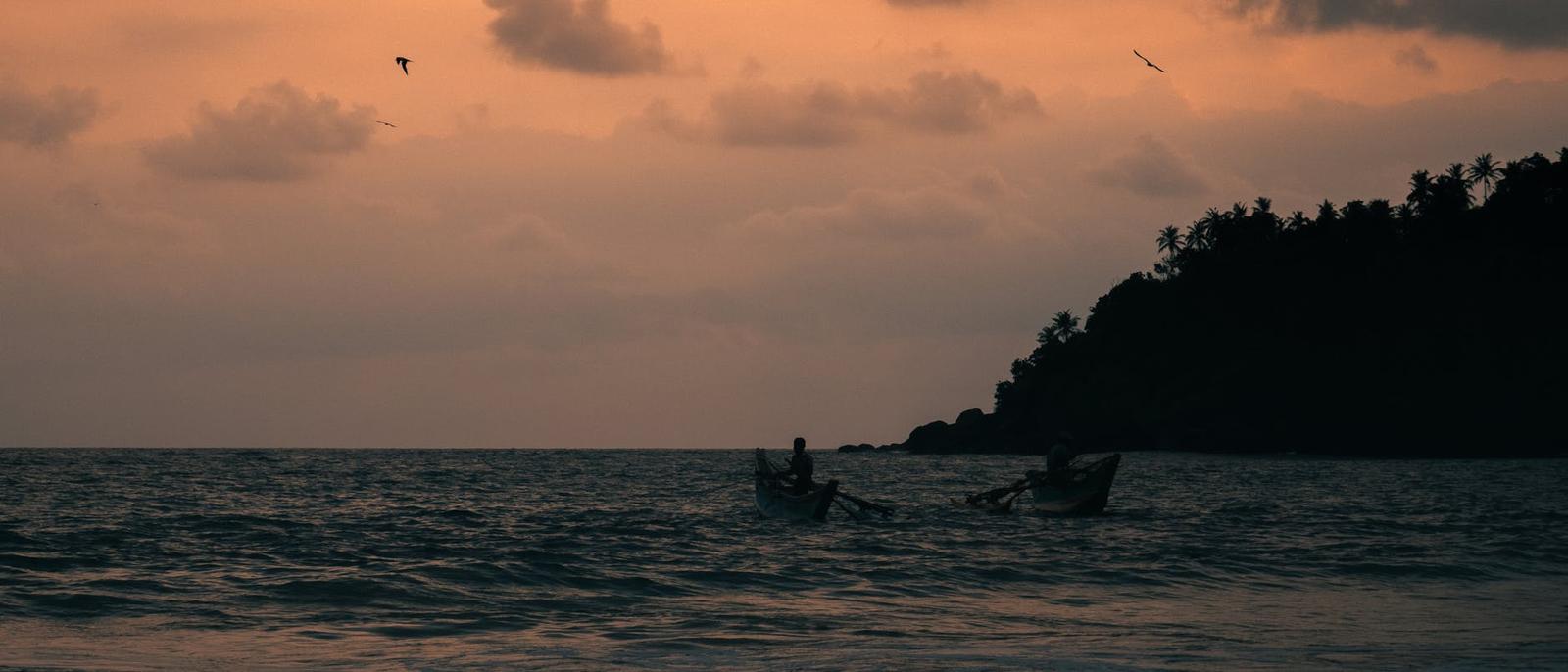 Two small fishing boats silhouetted against an orange sunset near a wooded headland