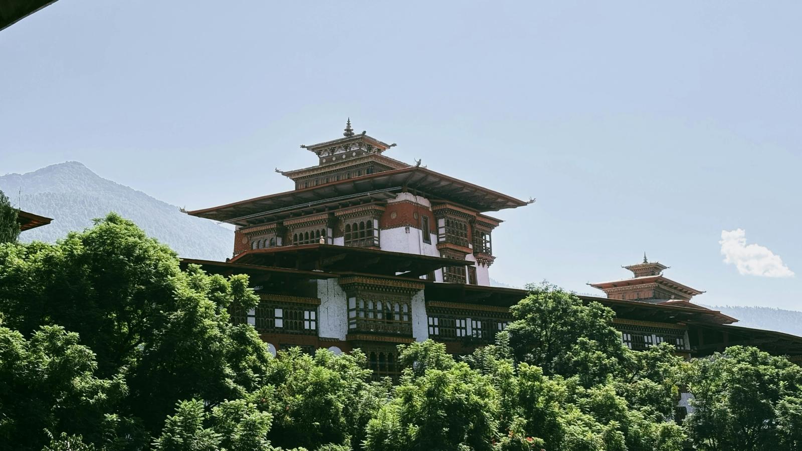 Traditional bhutanese architecture with ornate rooftops and trees. The grand Punakha Dzong rises above green treetops, its intricate Bhutanese architecture glowing under a bright sky with the Himalaya