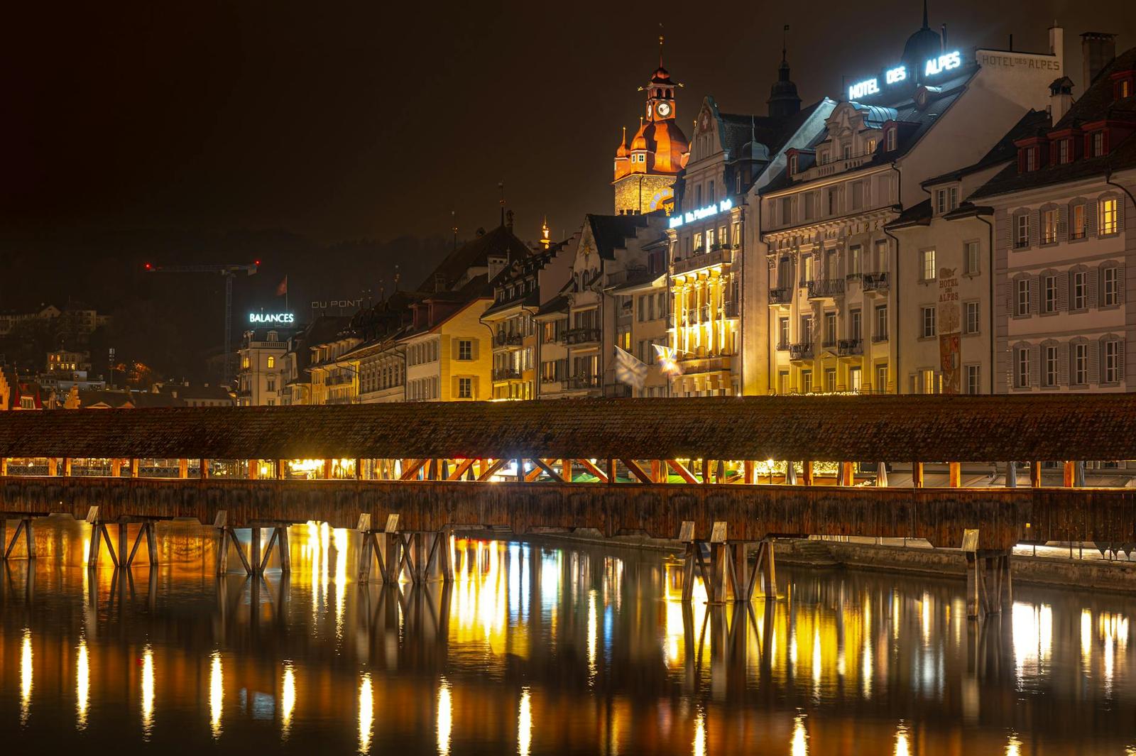 Wooden covered bridge crossing a river below a lit old town skyline at night