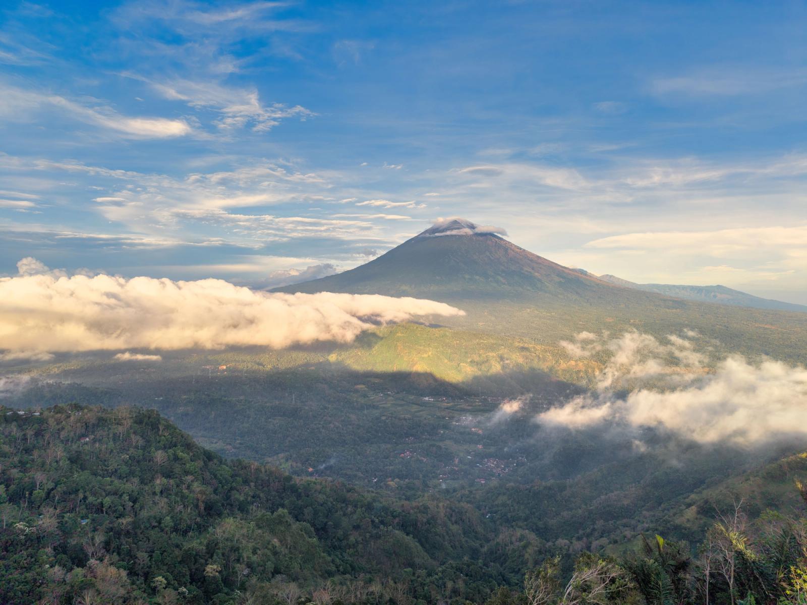 A smoking volcanic peak rising above forested hills wrapped in cloud