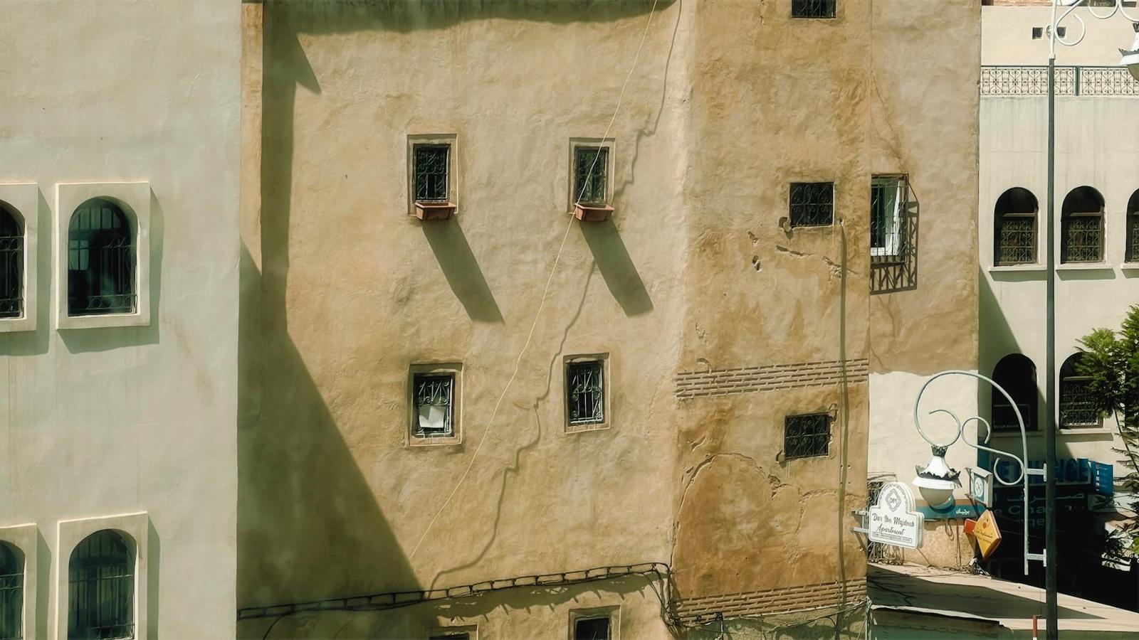 A weathered stucco building facade with small shuttered windows in sunlight