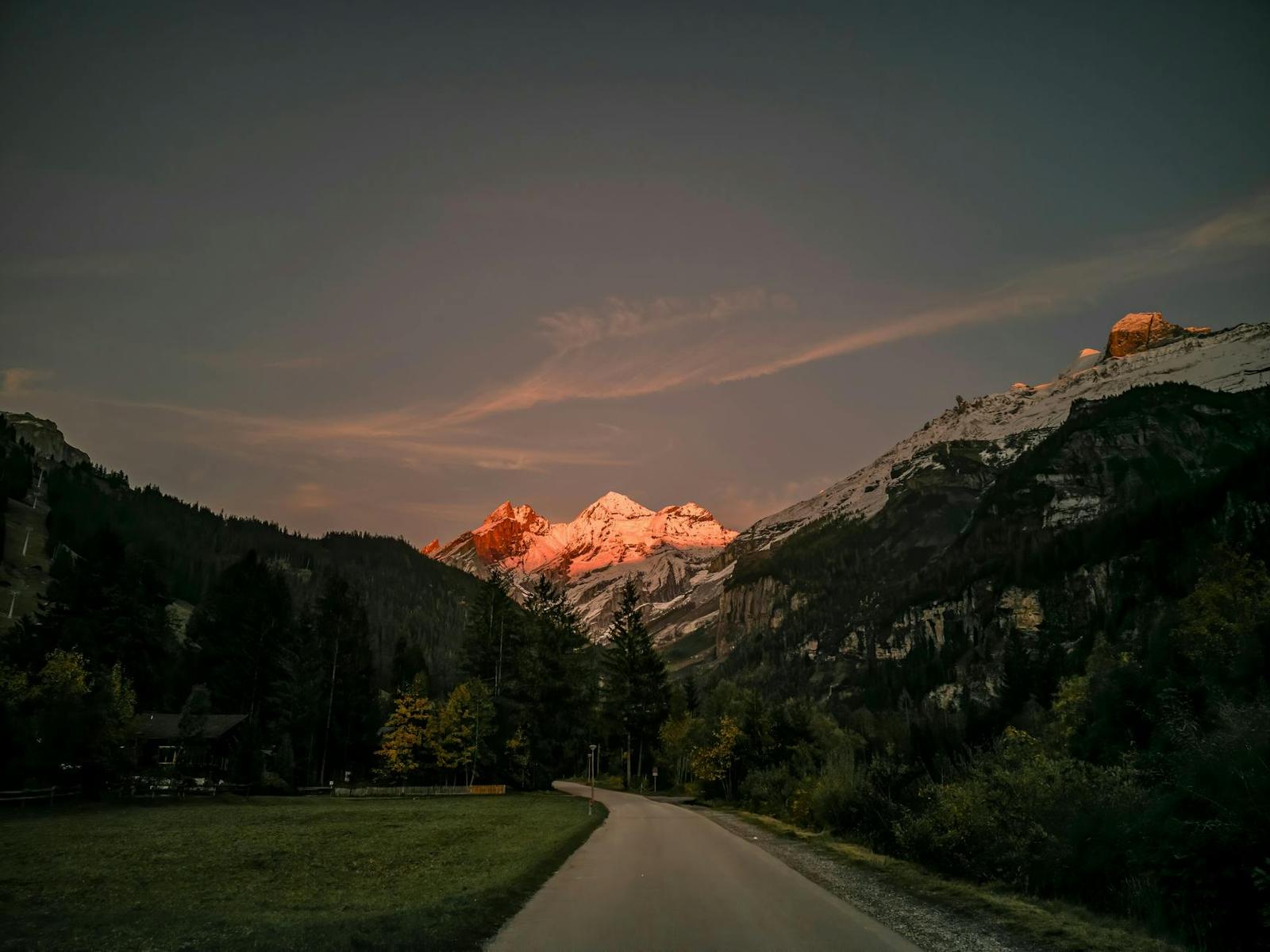 A quiet mountain road at dusk with snow peaks glowing pink above pine forest