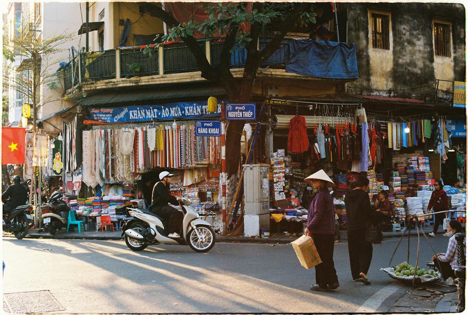 A busy street corner with market stalls, scooters and shoppers under a balcony