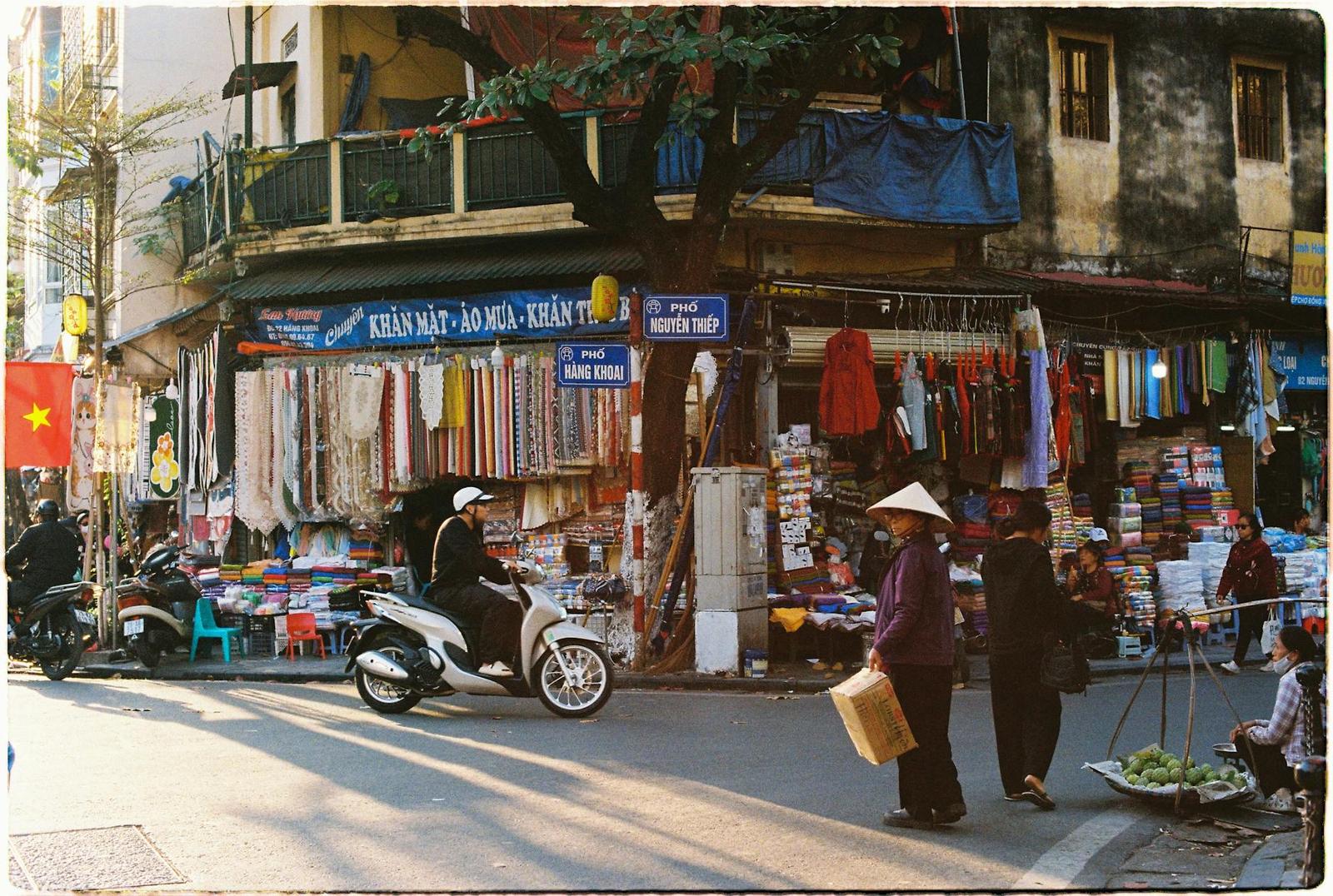A busy street corner with market stalls, scooters and shoppers under a balcony