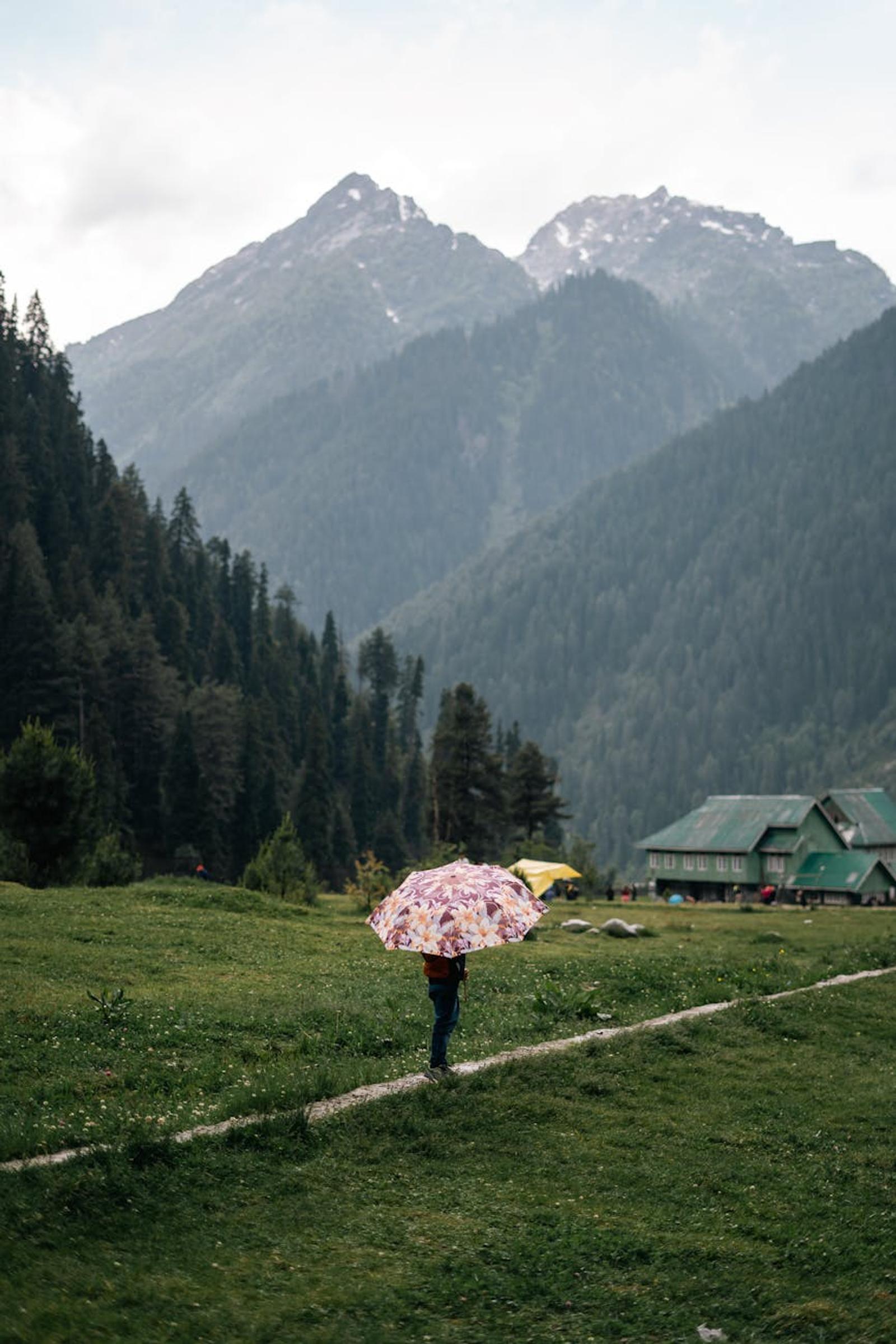 A person walks under an umbrella on a lush path in Pahalgam with mountains backdrop