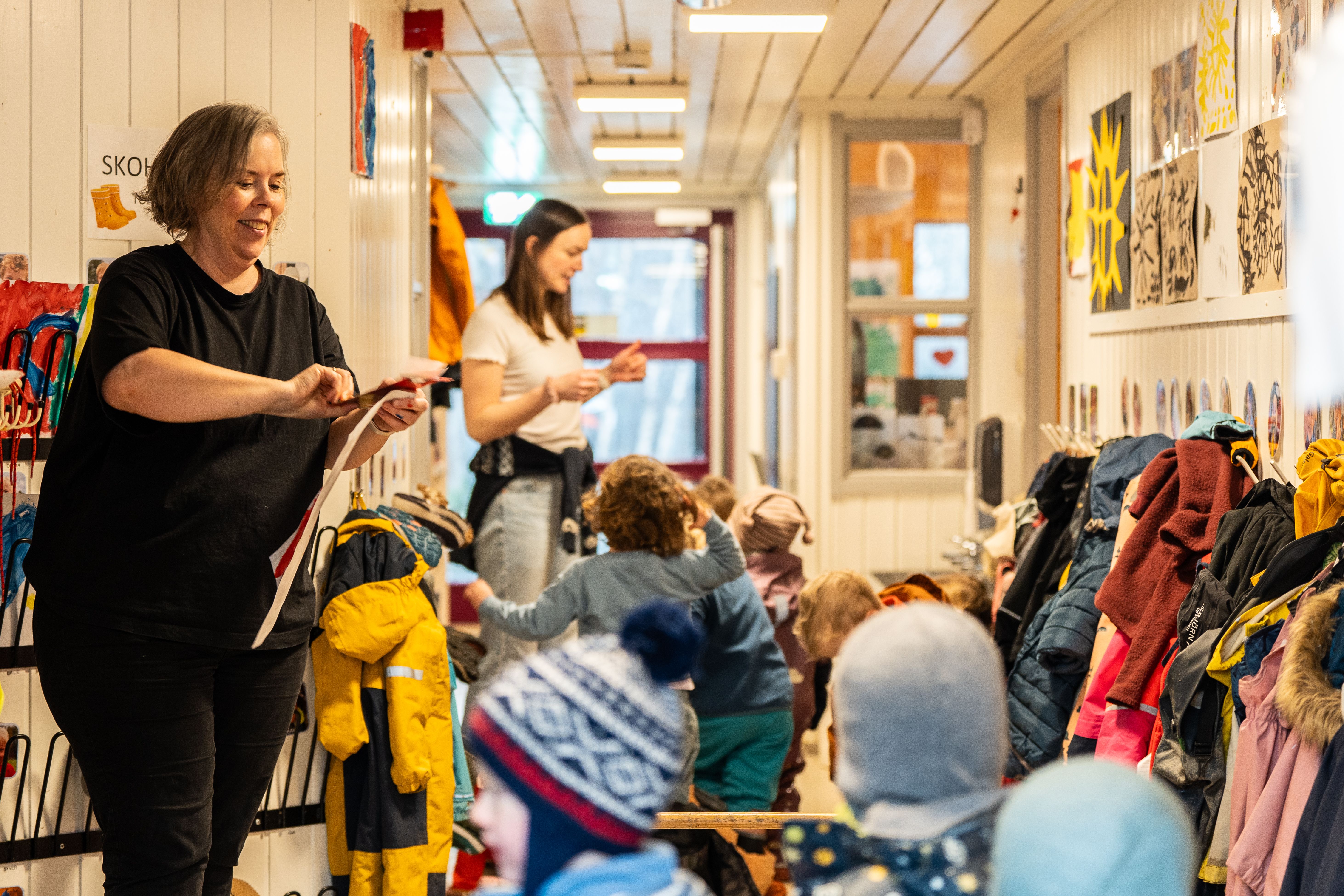 Foto av barnehageansatte som hjelper barna i garderoben med påkledning. Barn i ulike fargerike klær er i aktivitet, mens jakker og tegninger henger på veggene.