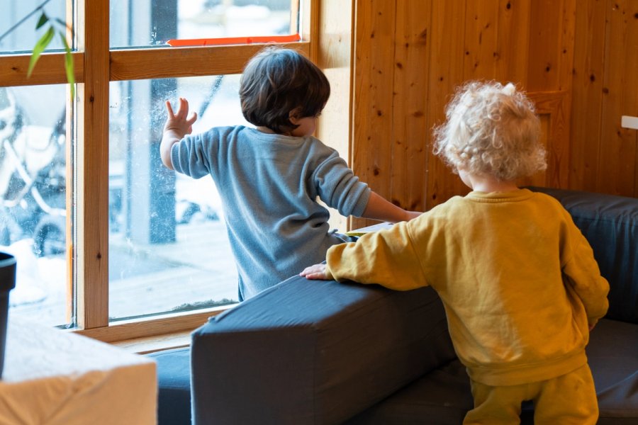 Two children playing by the window. One child touches the window. They are surrounded by cushions.
