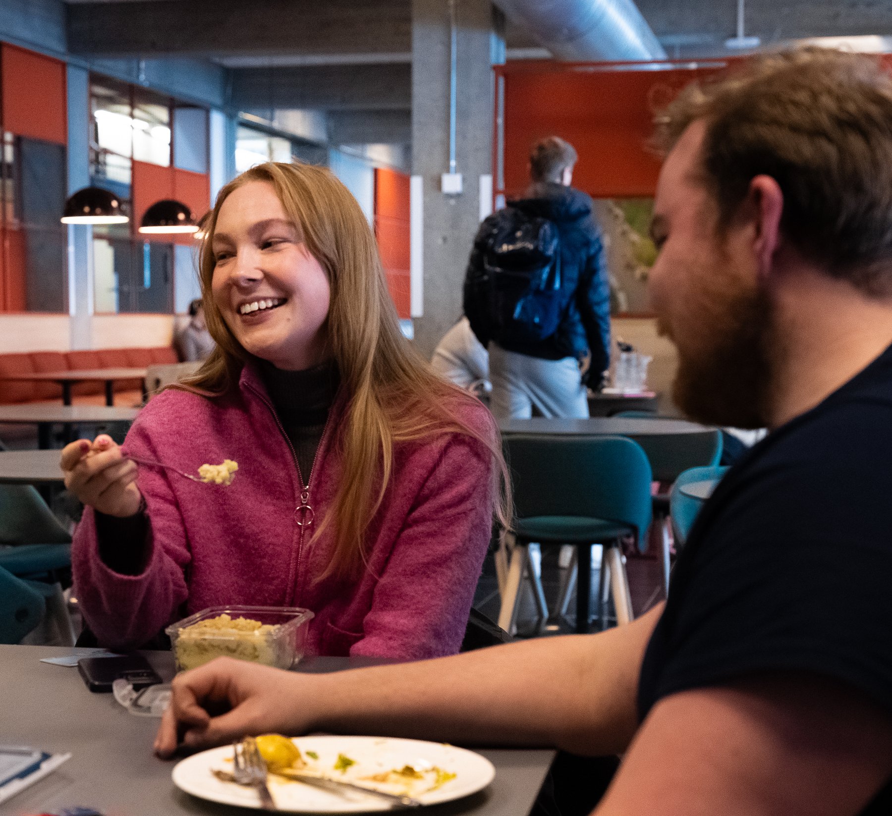 Two students sitting together in a canteen, eating and chatting.