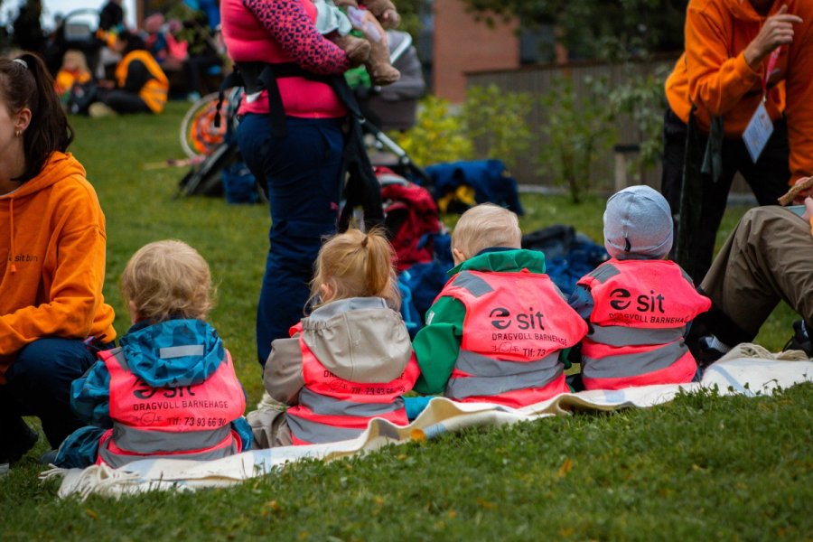 Fire små barn med rosa refleksvester fra Dragvoll barnehage sitter på et teppe i gresset, vendt mot en aktivitet under MiniUKA. Voksne og frivillige i oransje gensere er synlige i bakgrunnen.