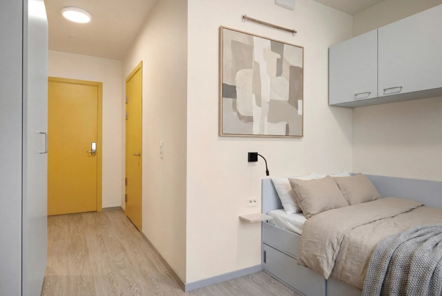 Bilde av en moderne studentbolig med seng, oppbevaringsløsninger, skrivepult og gang med inngangsdør i lyse farger.