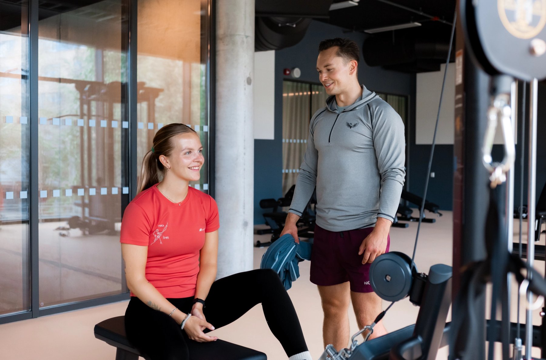 Photo of a man and woman in gym clothing smiling and talking by a bench inside the gym.