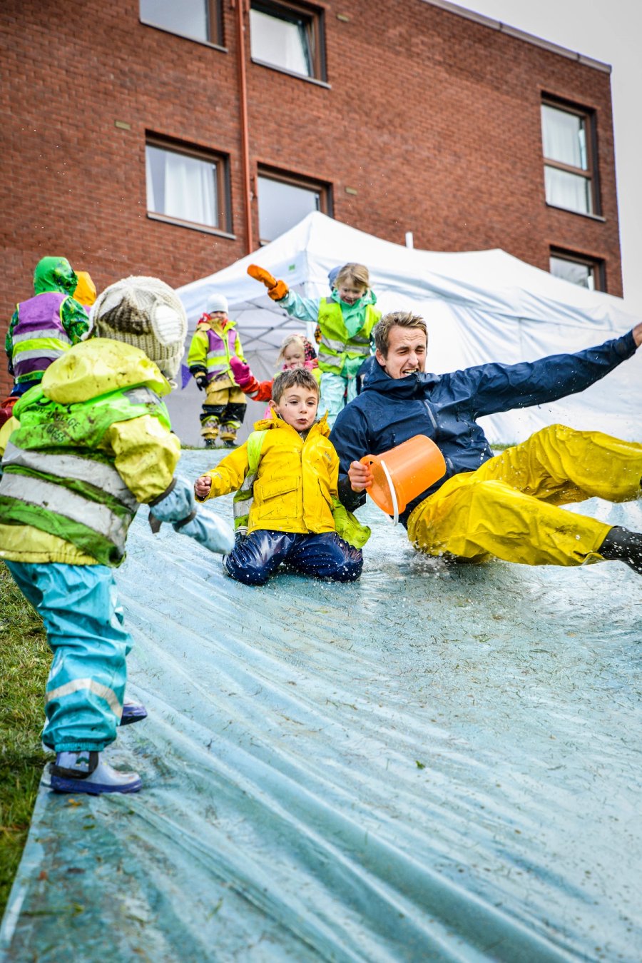 Barn og en voksen suser ned en våt plastsklie ute, iført regntøy og refleksvester. Vann spruter, og stemningen er preget av lek og entusiasme under MiniUKA.
