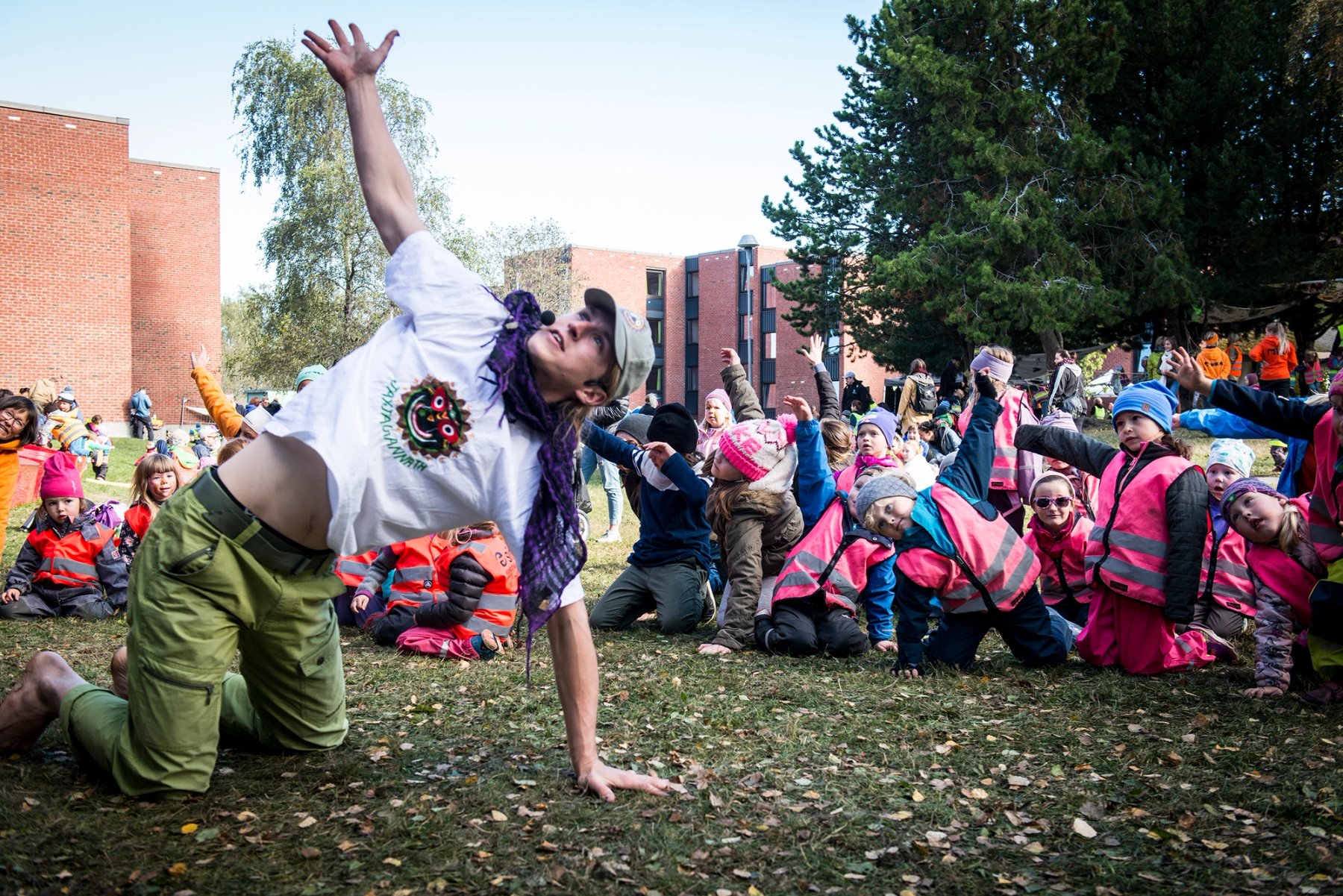 Et stort antall barn i refleksvester og voksne i oransje gensere sitter i sirkel på en gressplen, omgitt av høye trær og bygninger i bakgrunnen.