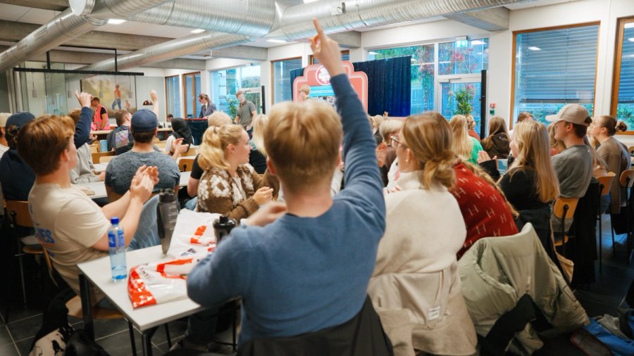 Studenter sitter samlet i et klasserom, mange med hendene i været under en quiz. Stemningen er engasjert og livlig.