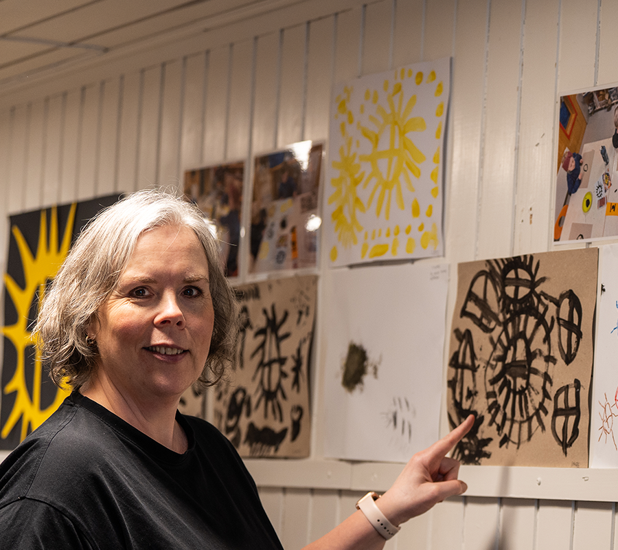 Photo of an educator who points to children’s artwork with sun motifs hanging on a white-painted wall. Drawings and paintings in bold colours are on display.