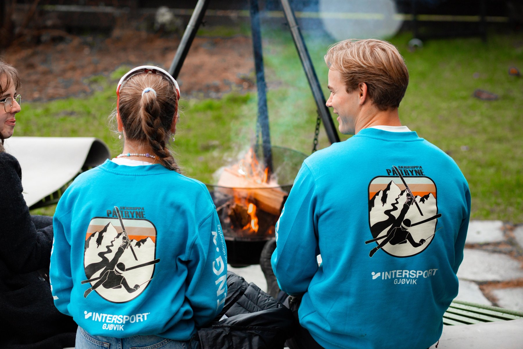 To studenter i like turkise gensere sitter ved et bål utendørs og snakker sammen.