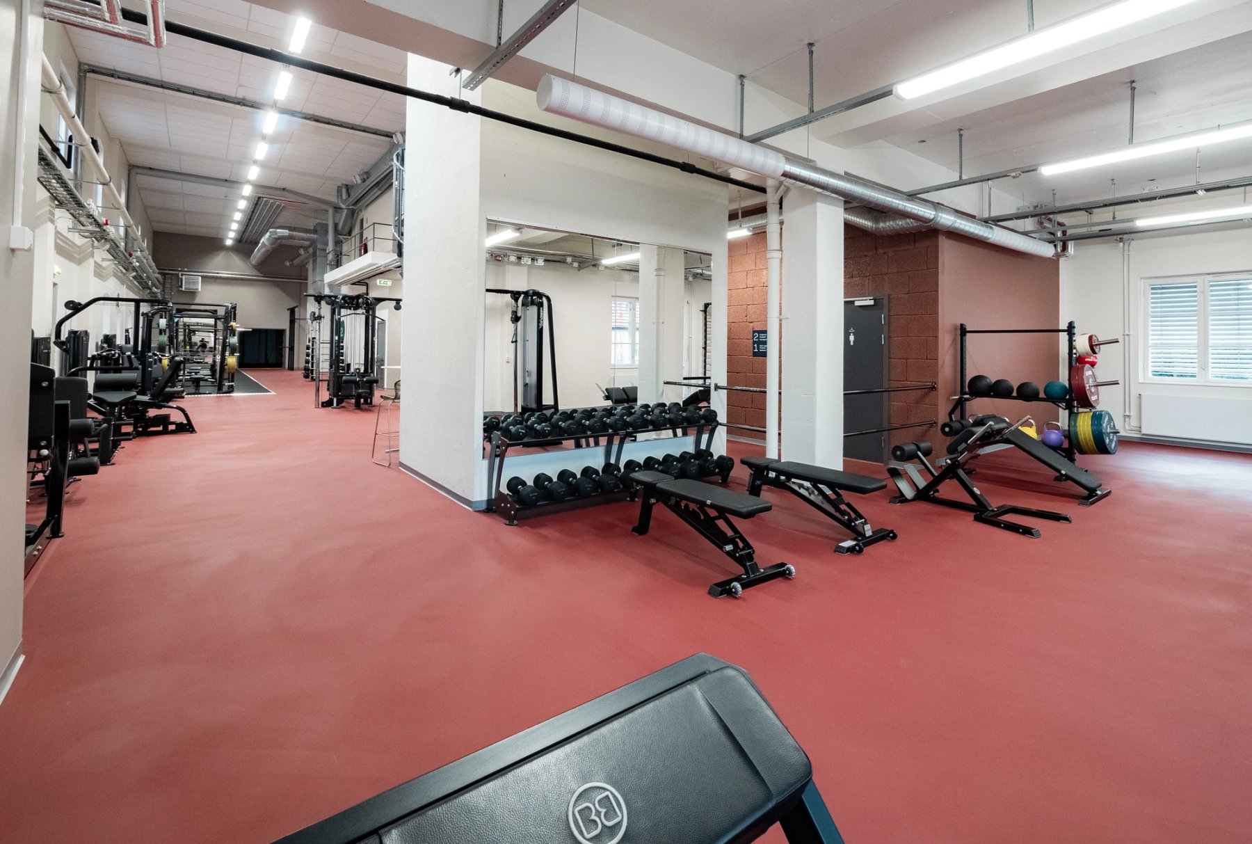 Oversiktsbilde over treningssenteret. Nærmest i bildet er noen treningsbenker og manualer foran et speil. Videre inn ser du magasinapparater og knebøystativ.