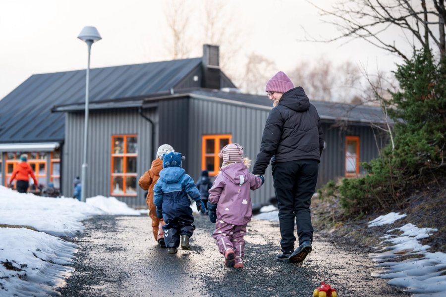 A kindergarten staff member walks hand in hand with children in winter suits along a snow-covered path outside the kindergarten building. More children and staff are walking ahead toward the building.